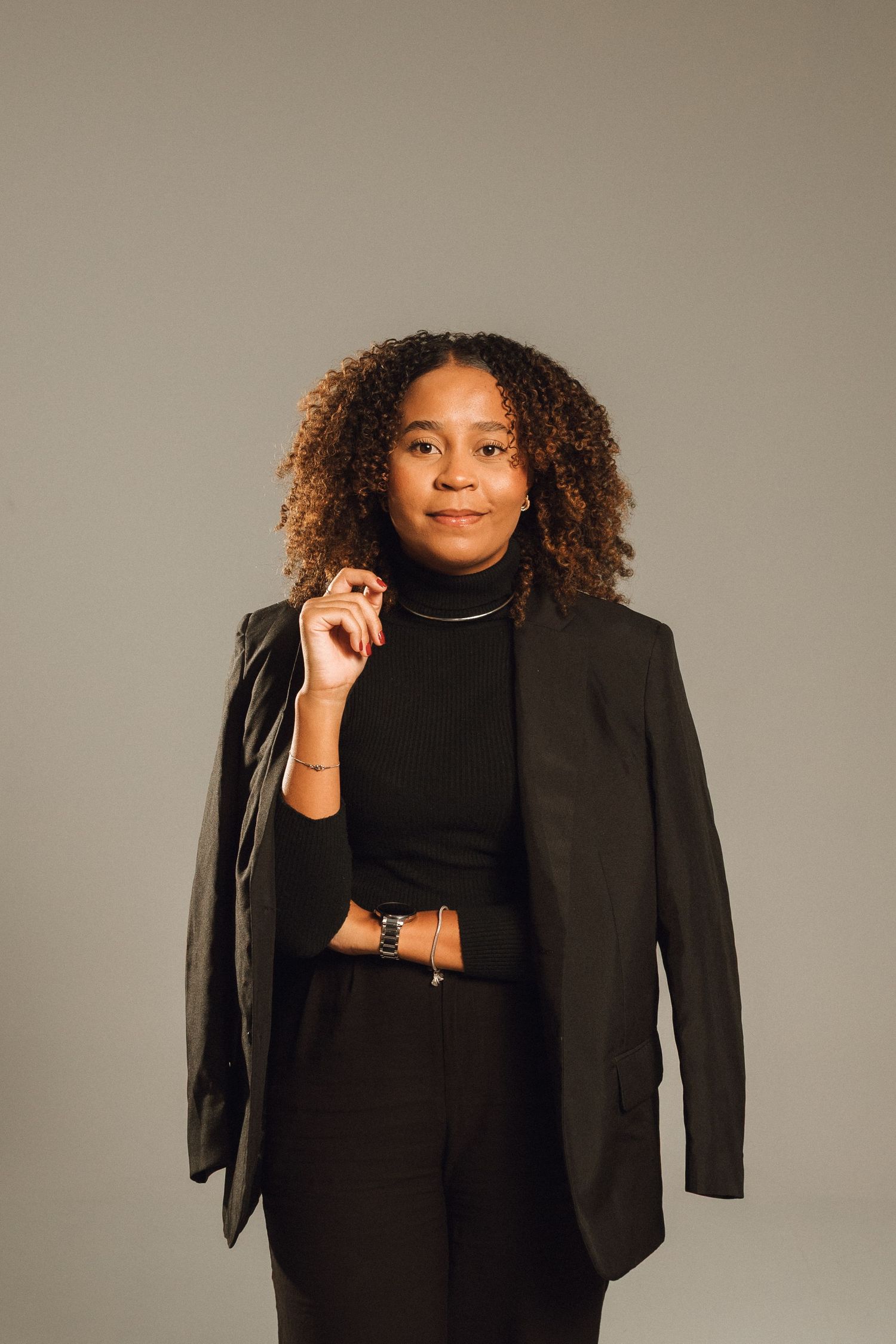 Confident Black woman in black turtleneck and blazer posing against neutral gray studio background.