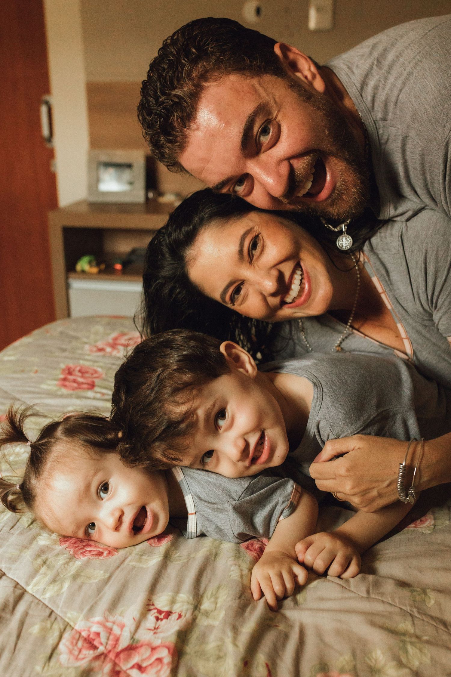 Happy family of four smiling together on a bed, with two young children and their parents in a cozy home setting.