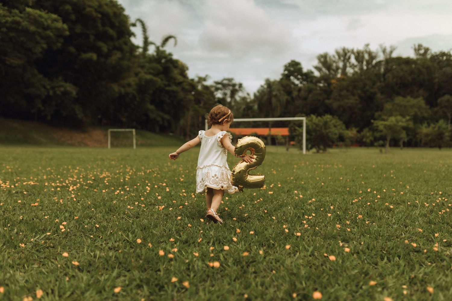 A toddler girl runs barefoot through a flower-filled green field holding a gold number 2 balloon on a cloudy day.