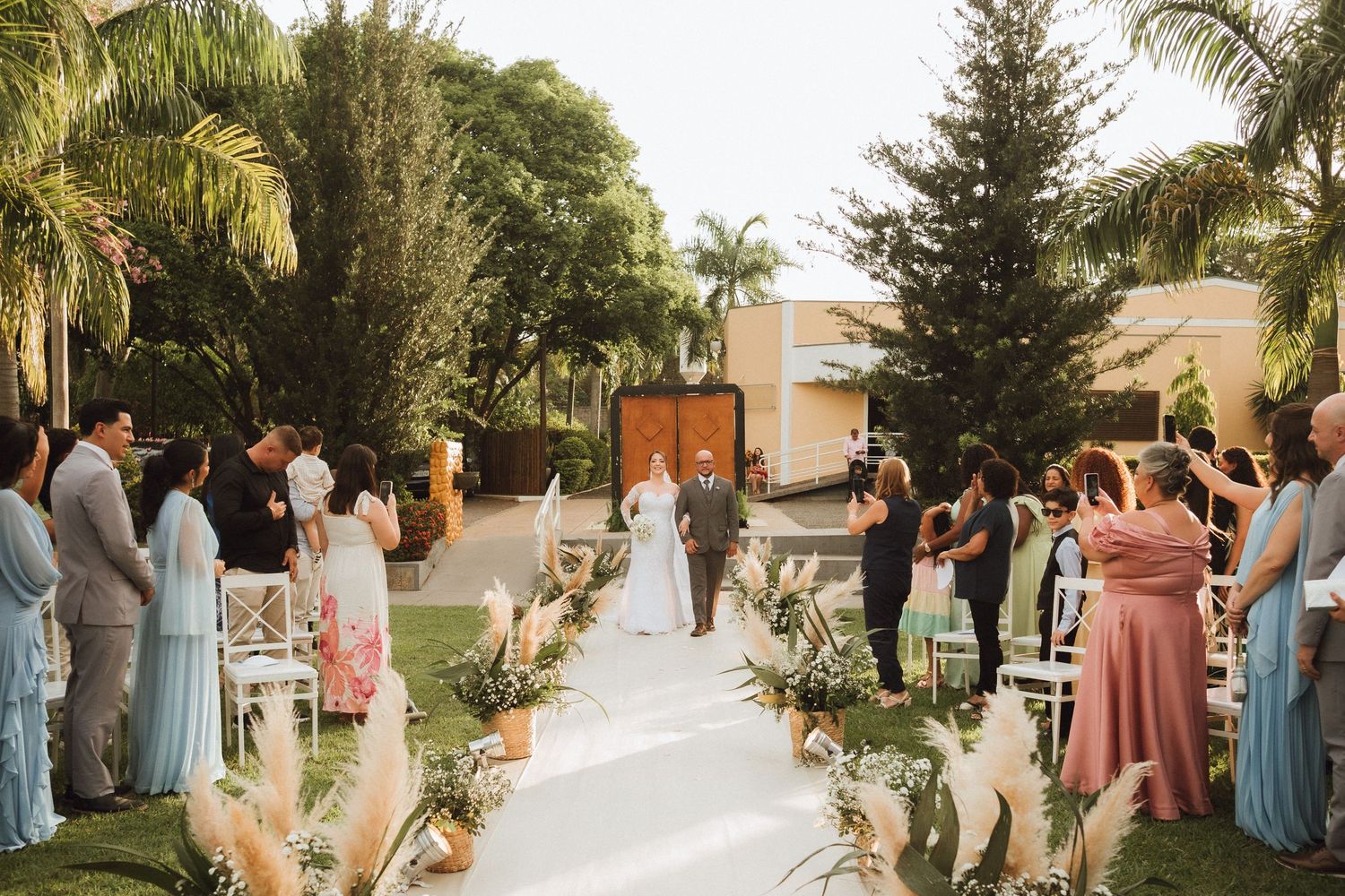 Outdoor garden wedding ceremony with white aisle, tropical trees, and guests in pastel attire.