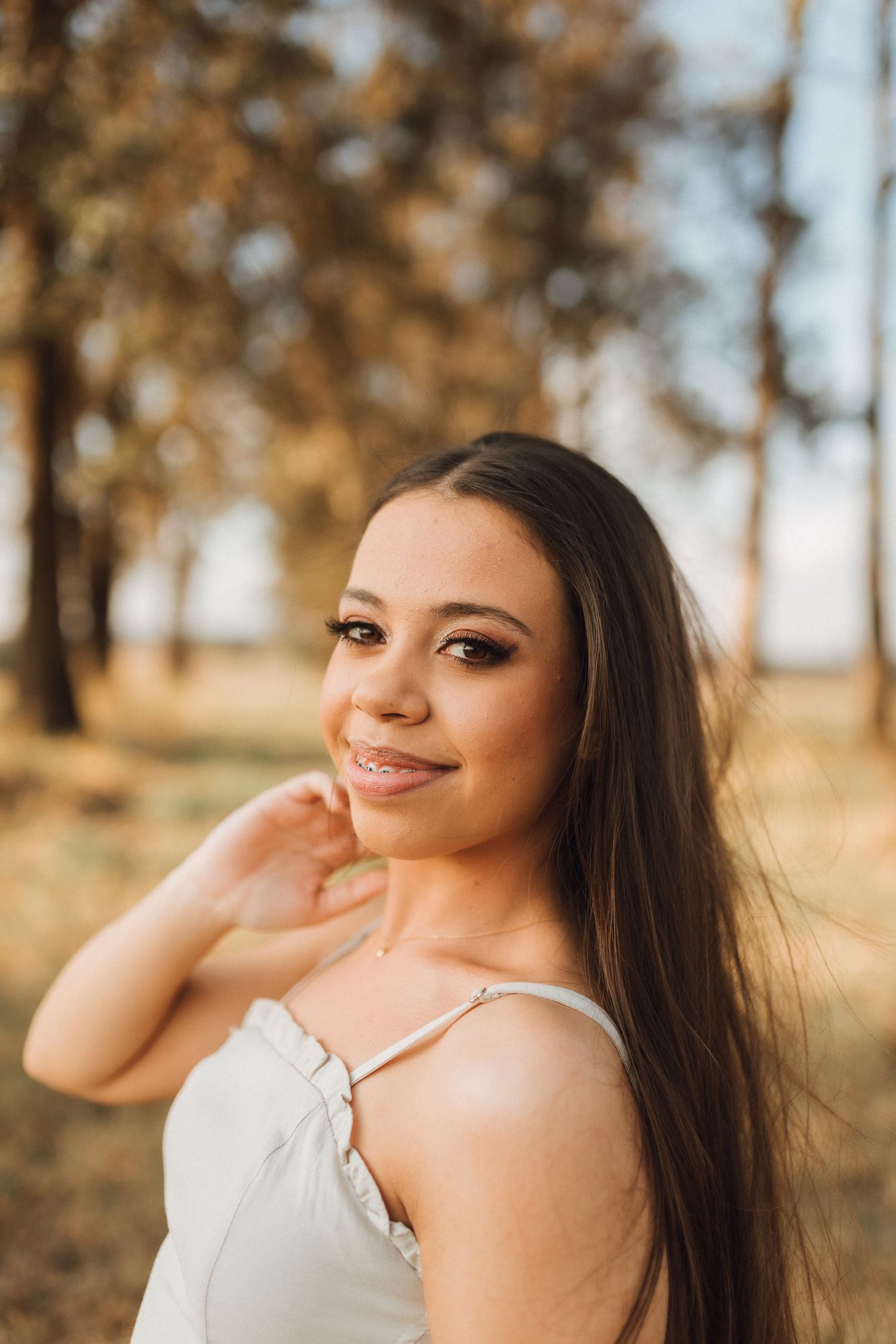 Young woman with long dark hair smiling outdoors in a white dress, surrounded by autumn trees in golden light.
