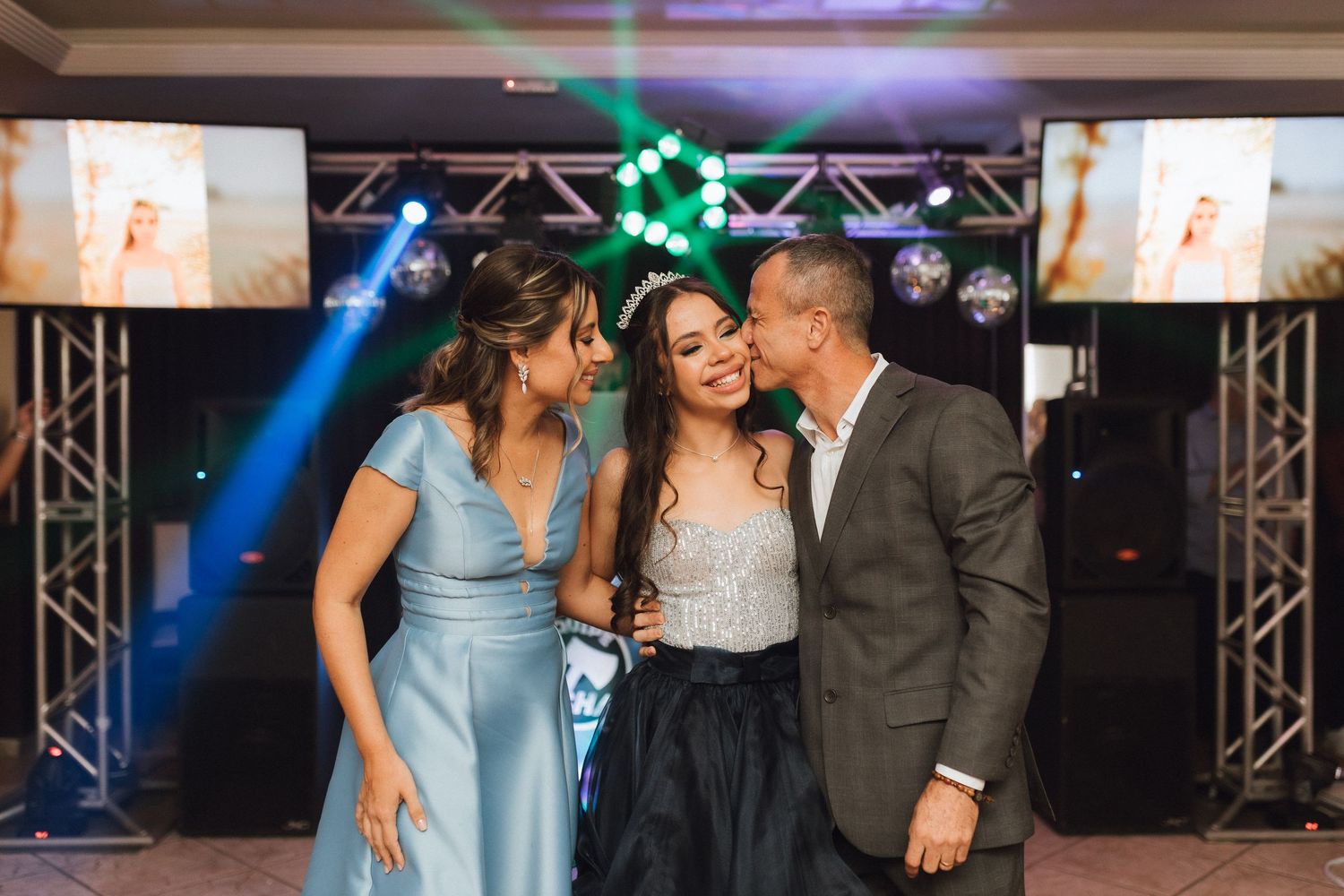 Parents kissing their daughter in tiara and black gown on LED dance floor at quinceañera party.