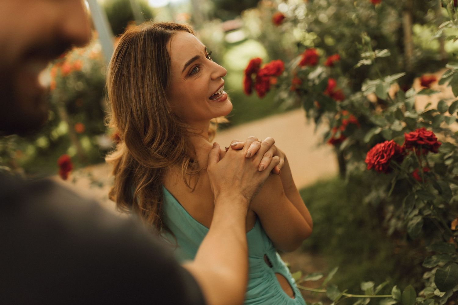 A smiling woman in a teal dress holds hands with a man near blooming red roses in a garden.