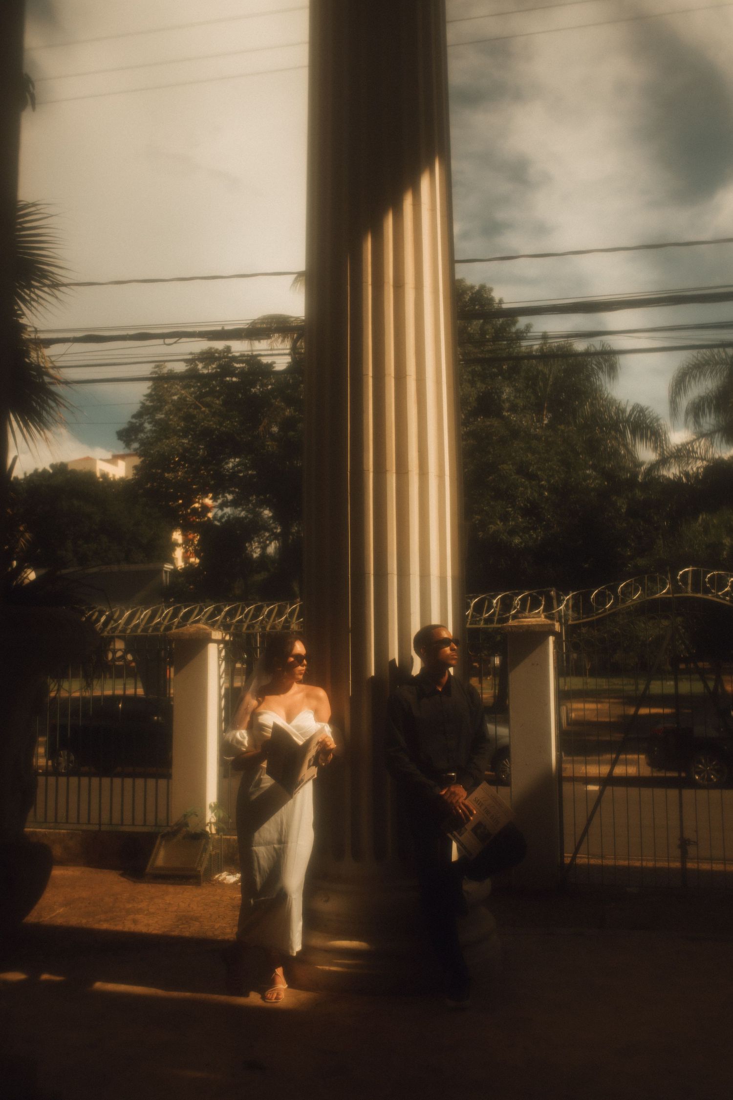 Couple posing by a grand column near ornate iron gates, surrounded by lush trees in warm sunlight.