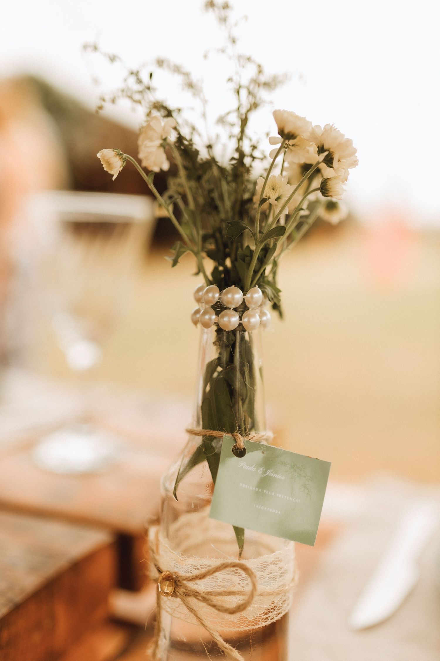 Rustic wedding table centerpiece with wildflowers in a glass vase wrapped in lace, twine, and pearl beads with a small card.