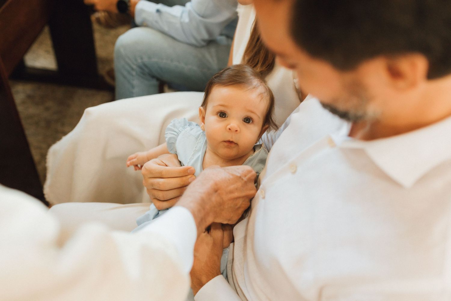 A baby in a light blue outfit being held by a bearded man in a white shirt during a baptism ceremony.