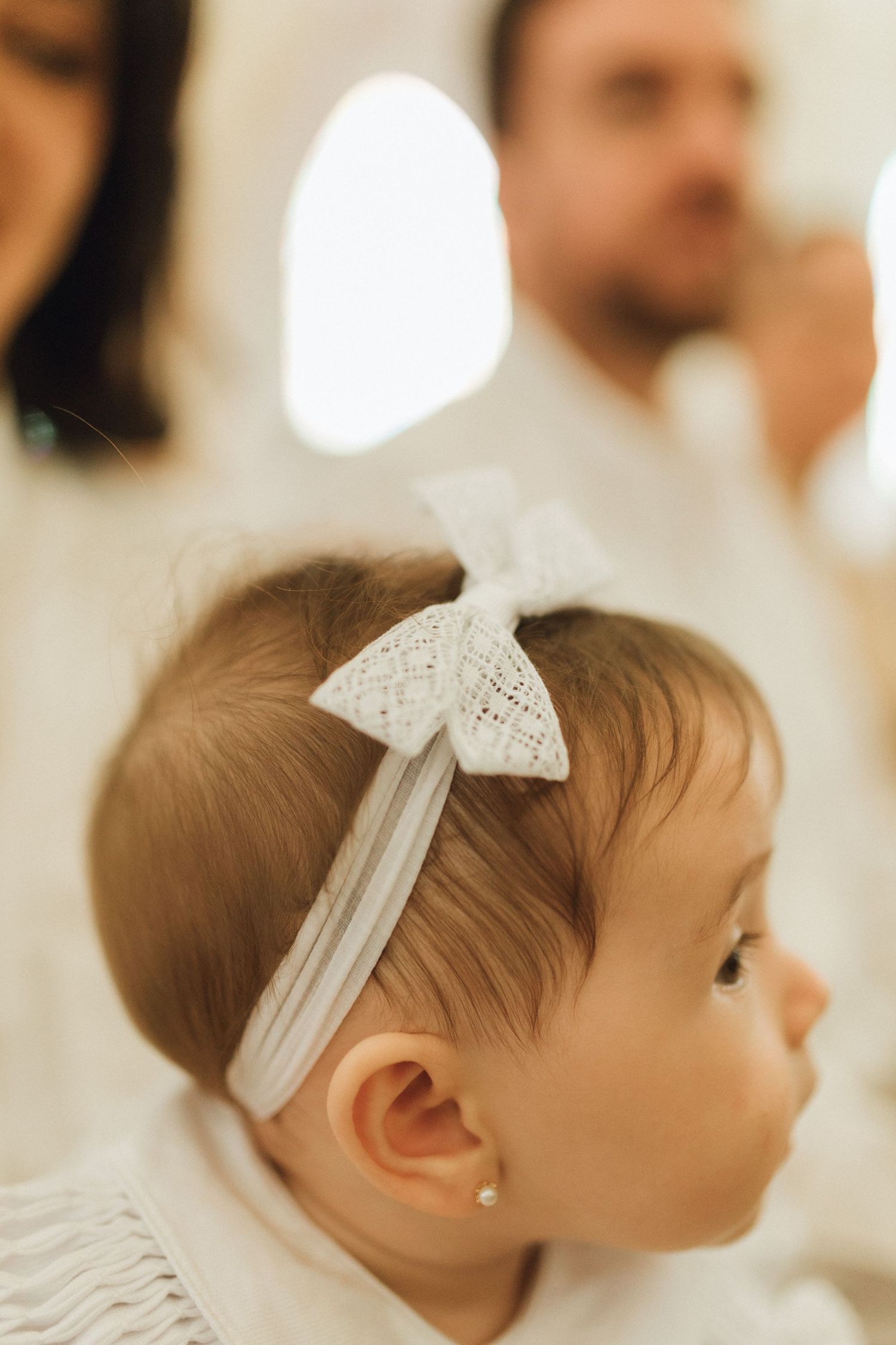 Close-up of a baby girl wearing a white lace bow headband and pearl earring at a christening ceremony.