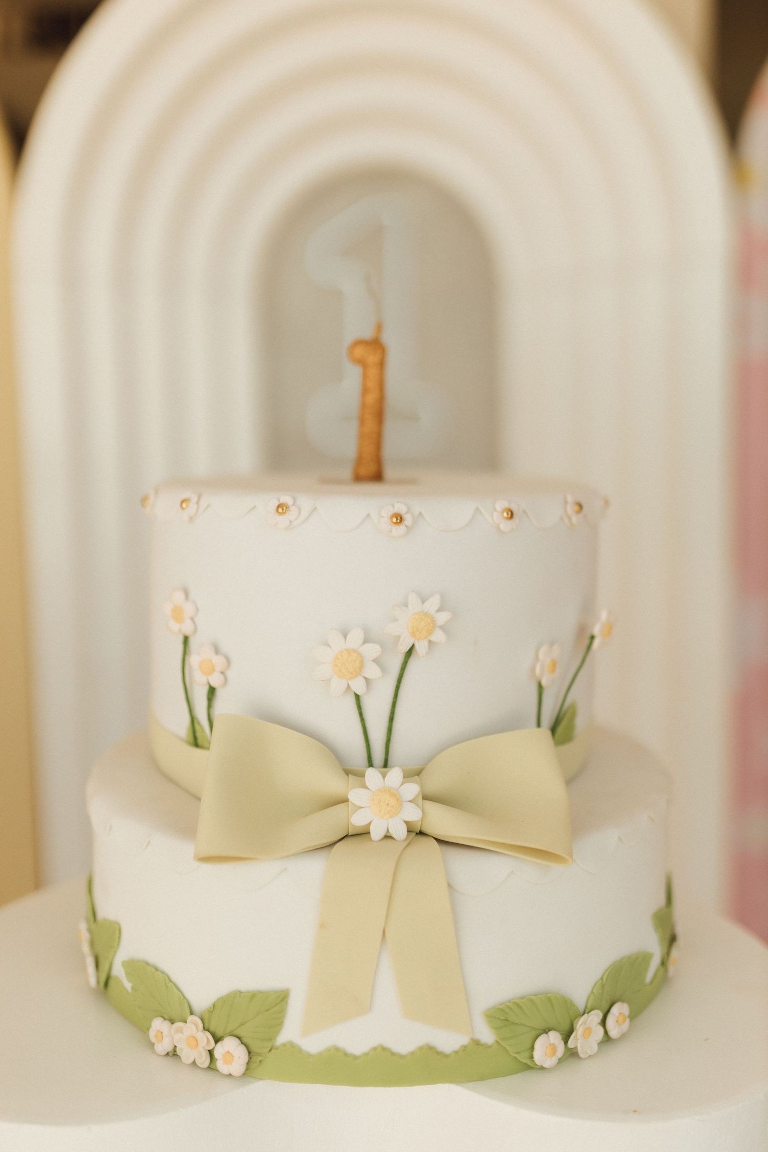 Two-tier white fondant birthday cake with daisy flowers, green bow, and number one candle topper.
