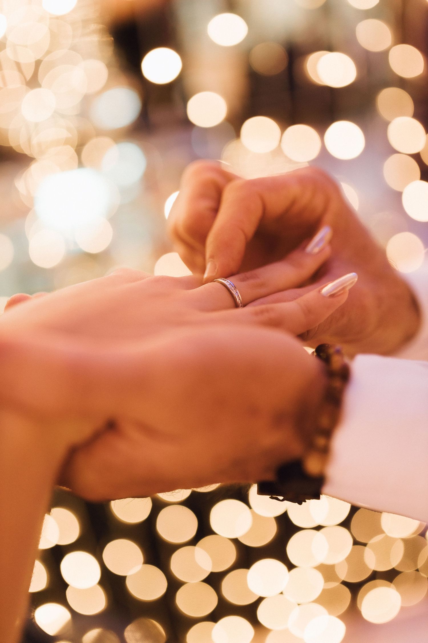 Close-up of groom placing wedding ring on bride's finger with golden bokeh lights in background.