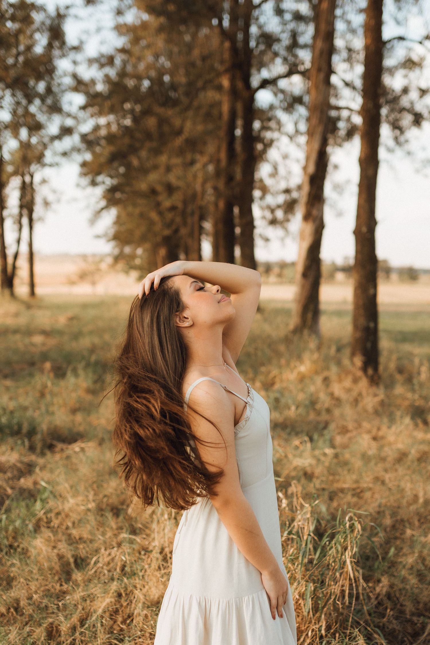 Young woman in white sundress posing gracefully in golden sunlit forest, flowing brown hair, eyes closed, serene nature setting.
