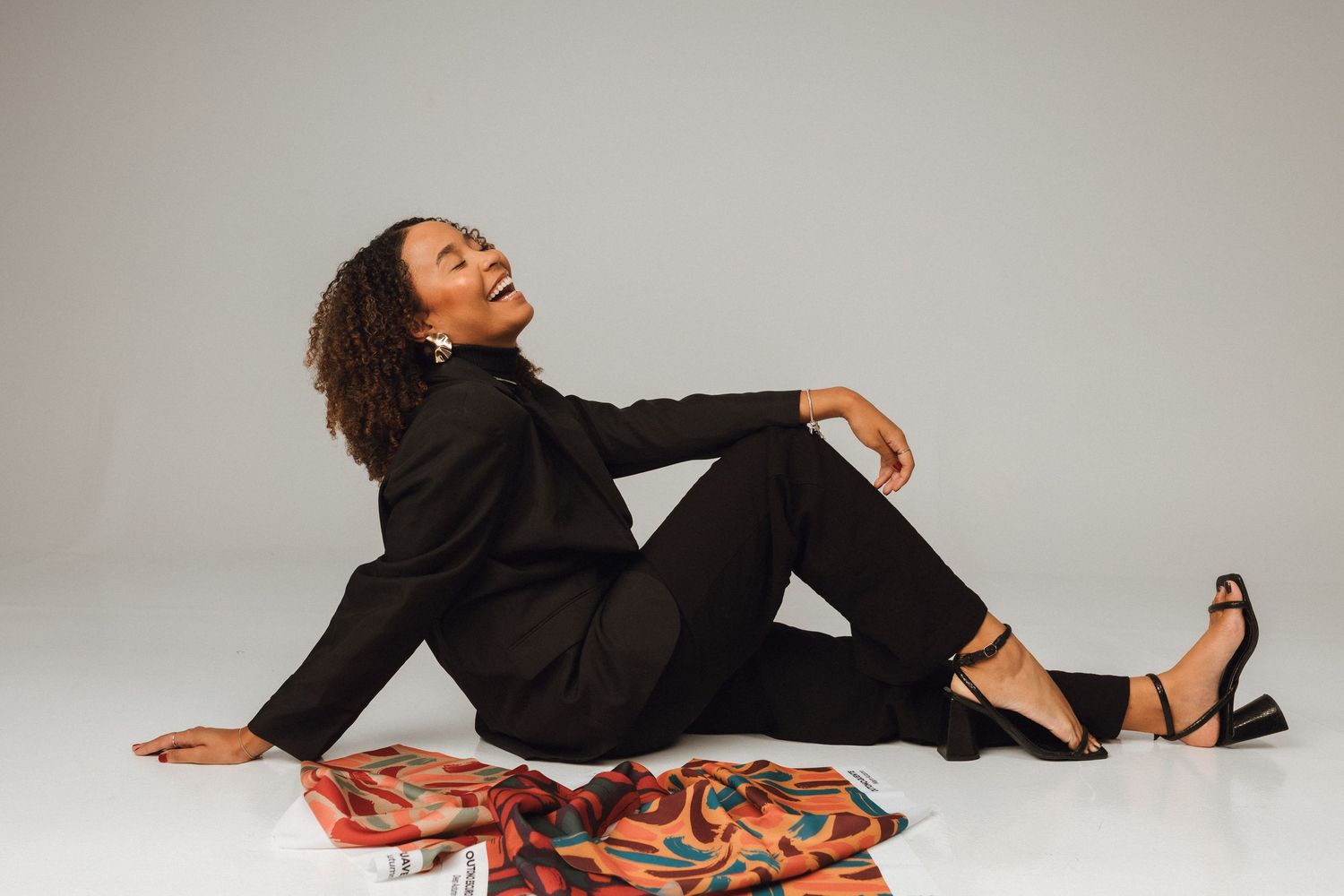 Woman in black suit sitting gracefully on floor beside colorful fabric in a professional studio setting.