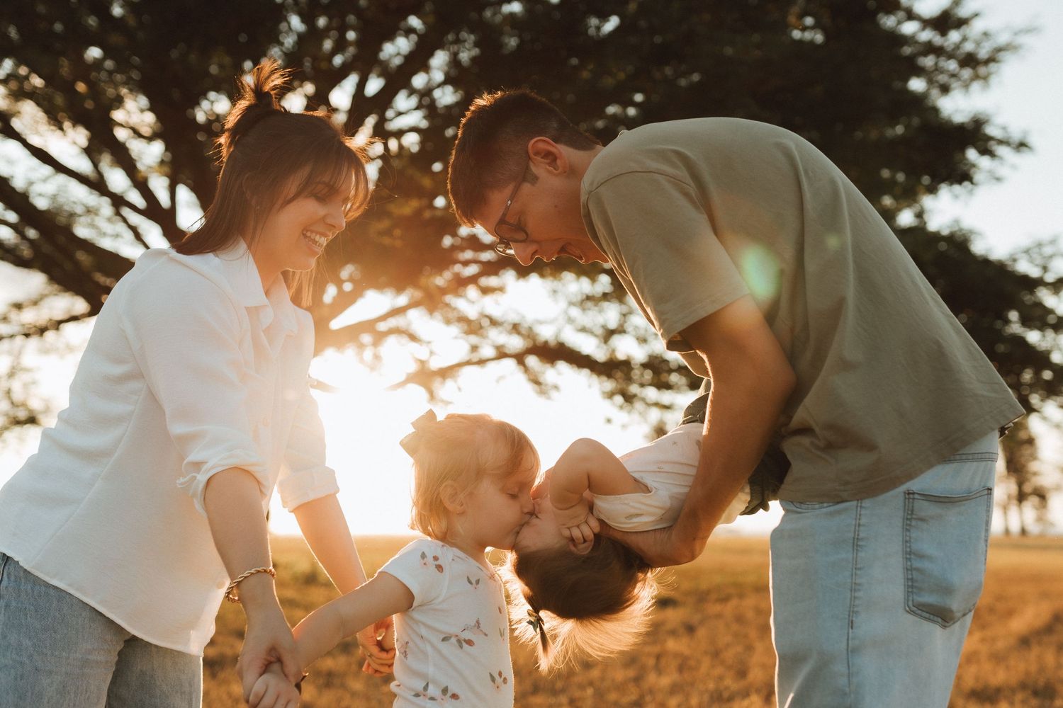 Parents laughing and playing with toddler in a glowing sunset field near a tree.