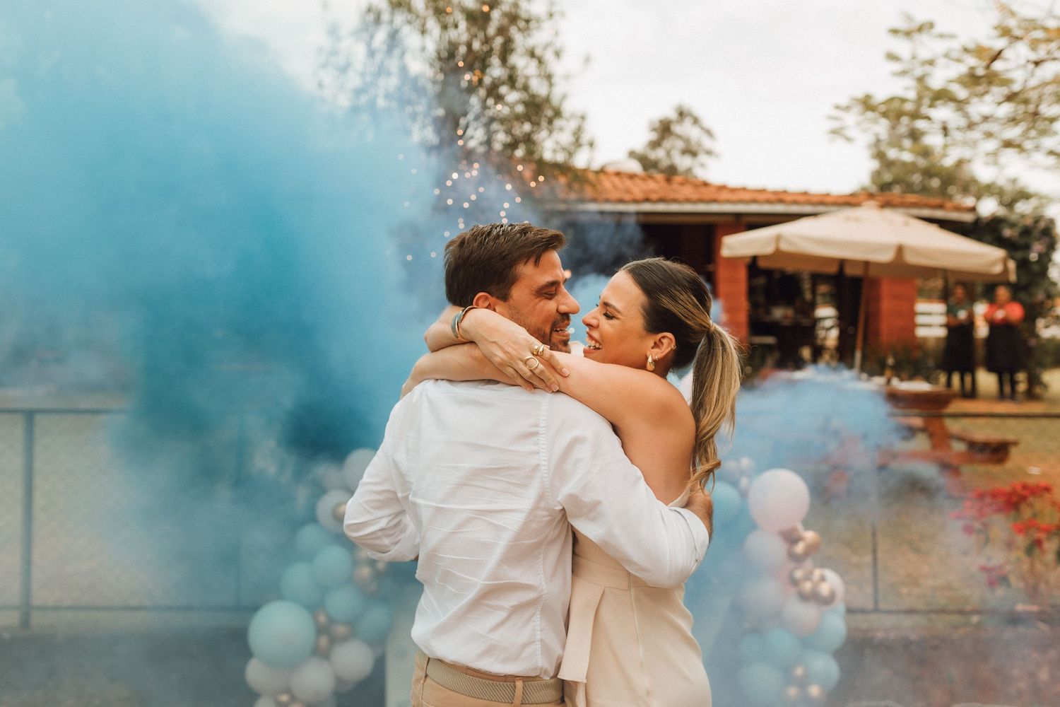 Couple embracing during gender reveal with blue smoke bombs and balloon arch at outdoor celebration.