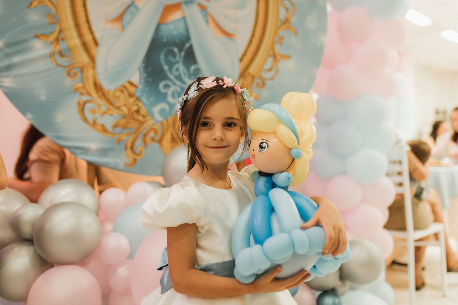 Young girl in white dress holds Cinderella doll at princess-themed birthday party with pink and blue balloons.