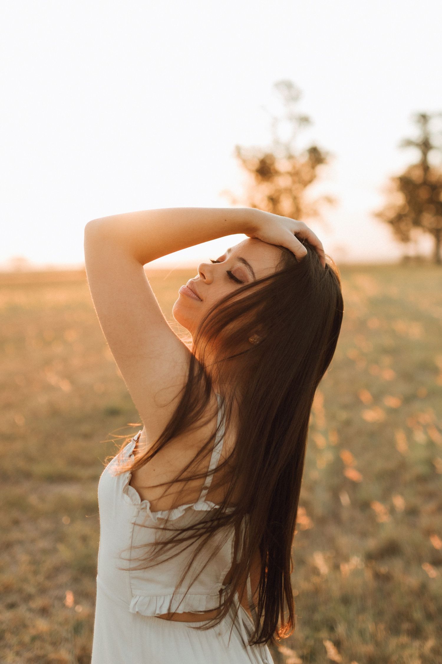 Young woman with long dark hair poses in golden sunset light in an open field, wearing a white dress.