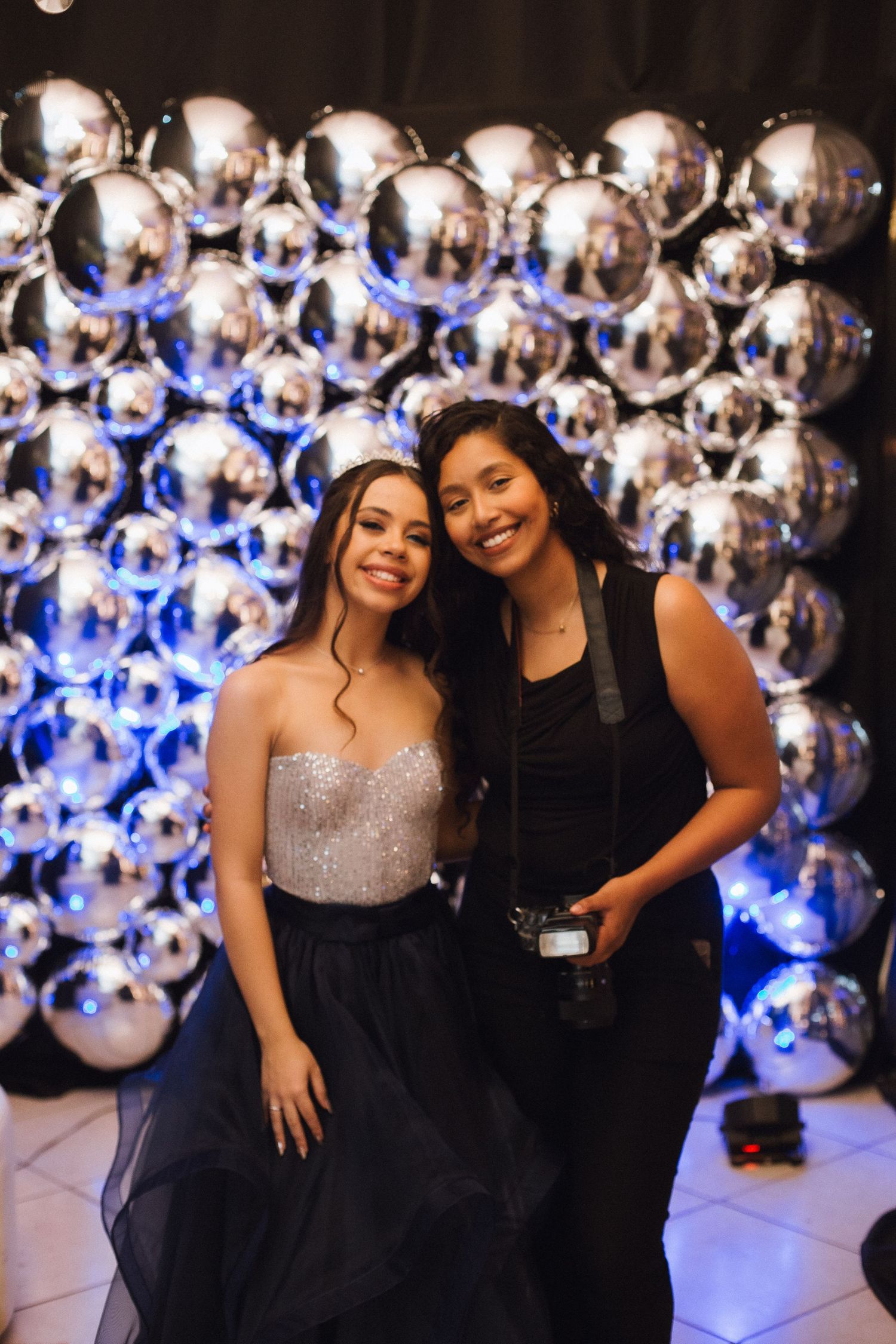 Two smiling women pose together in front of a shimmering silver balloon wall at a formal event.