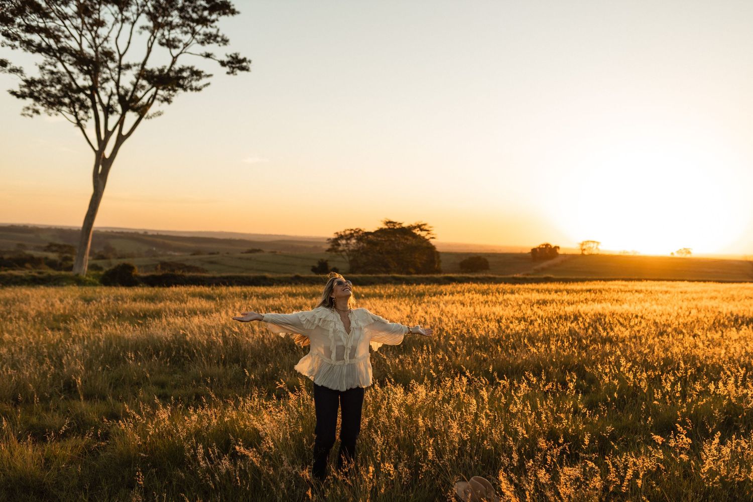 Woman standing in golden wheat field at sunset with arms outstretched, large tree silhouetted against warm orange sky.