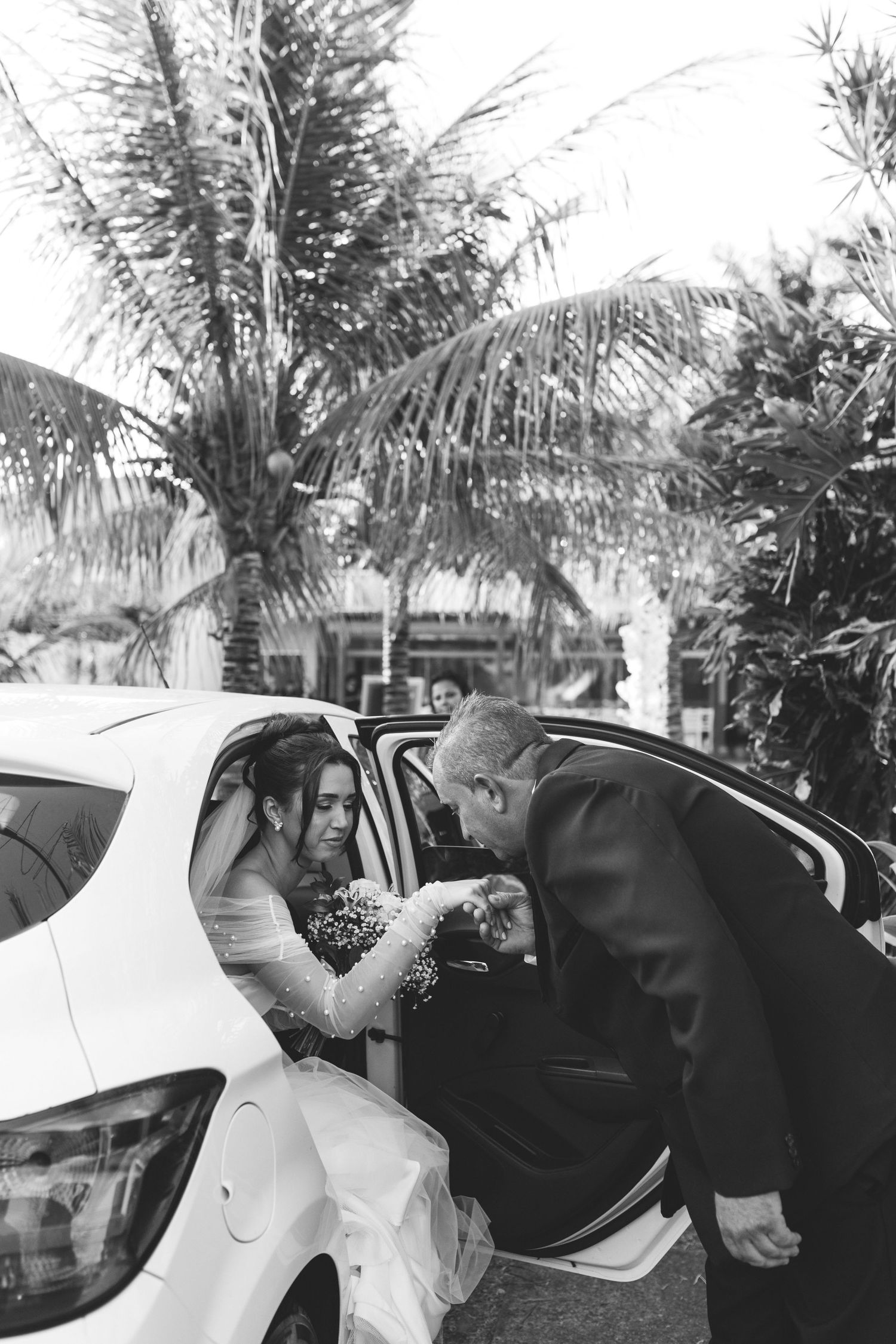 Black and white photo of groom helping bride exit a white car at outdoor wedding venue.