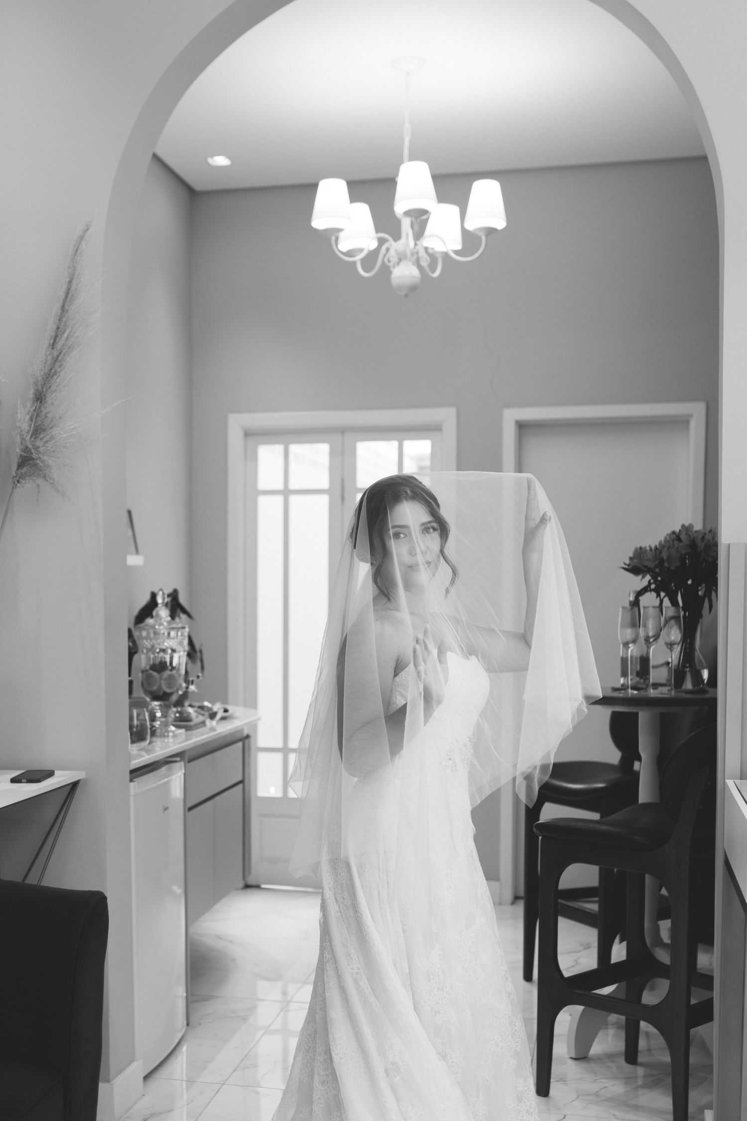 Black and white portrait of a bride in wedding dress and veil standing under a chandelier.