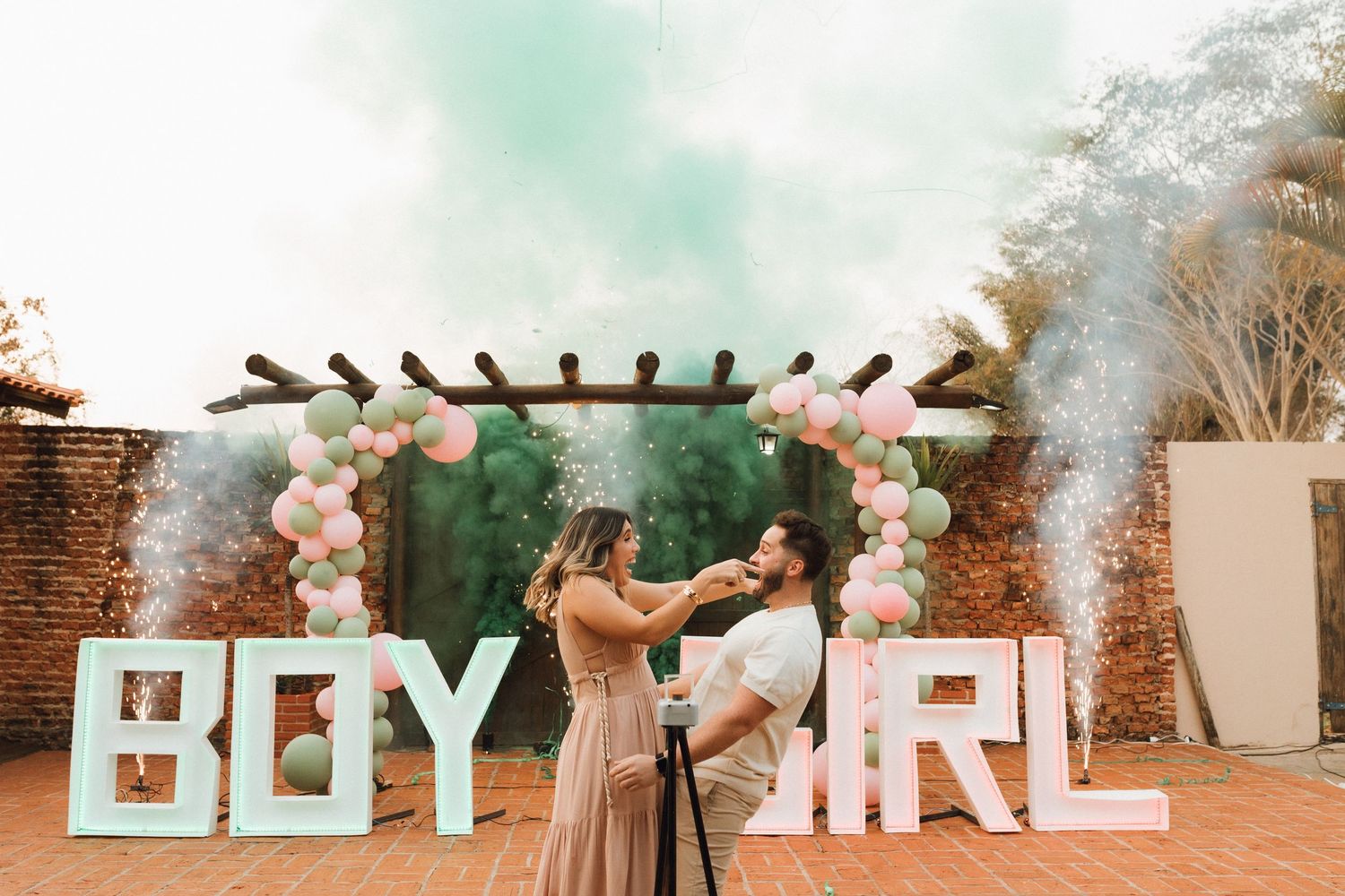 Couple celebrating gender reveal party with pink and green smoke bombs beside BOY and GIRL signs under balloon arch.