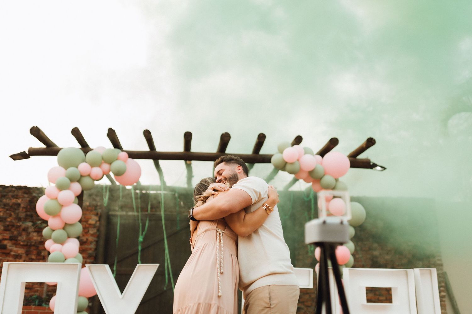 Couple embracing at gender reveal party with green smoke, pink and mint balloons, and decorative letters outdoors.