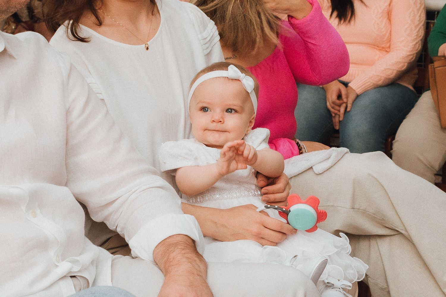 Adorable baby girl in white dress sitting on father's lap, holding colorful toy, with family gathered around.