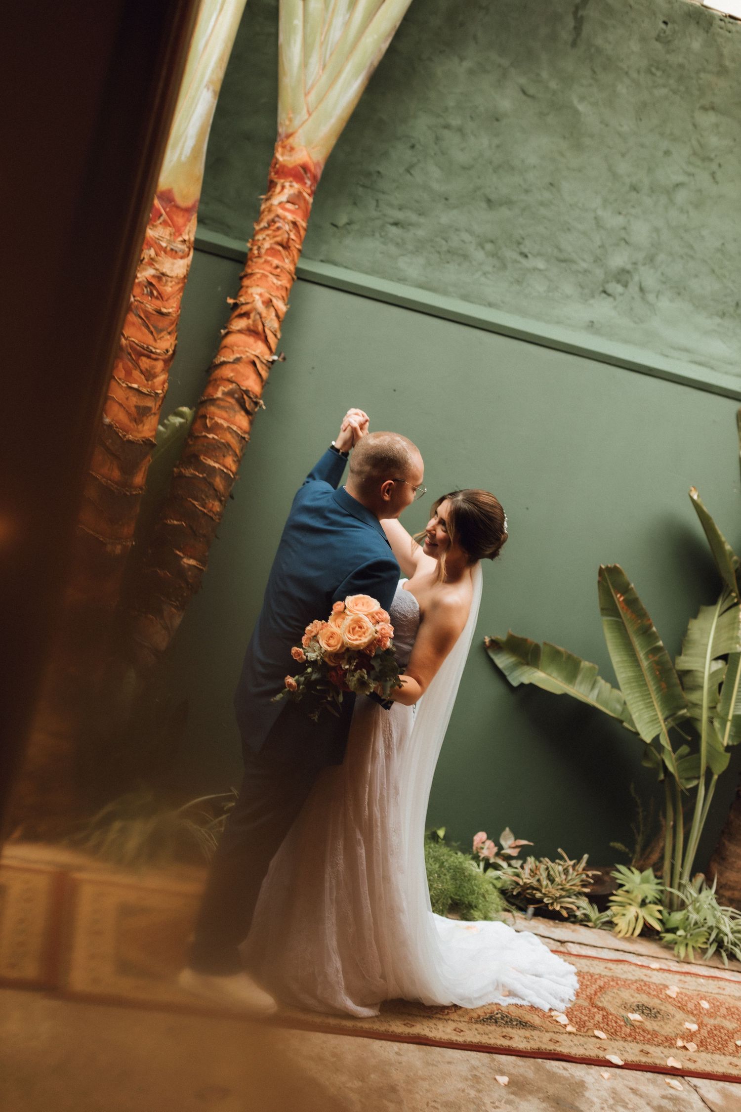 Groom dips bride in romantic first dance pose near tropical palm trees in green-walled venue.