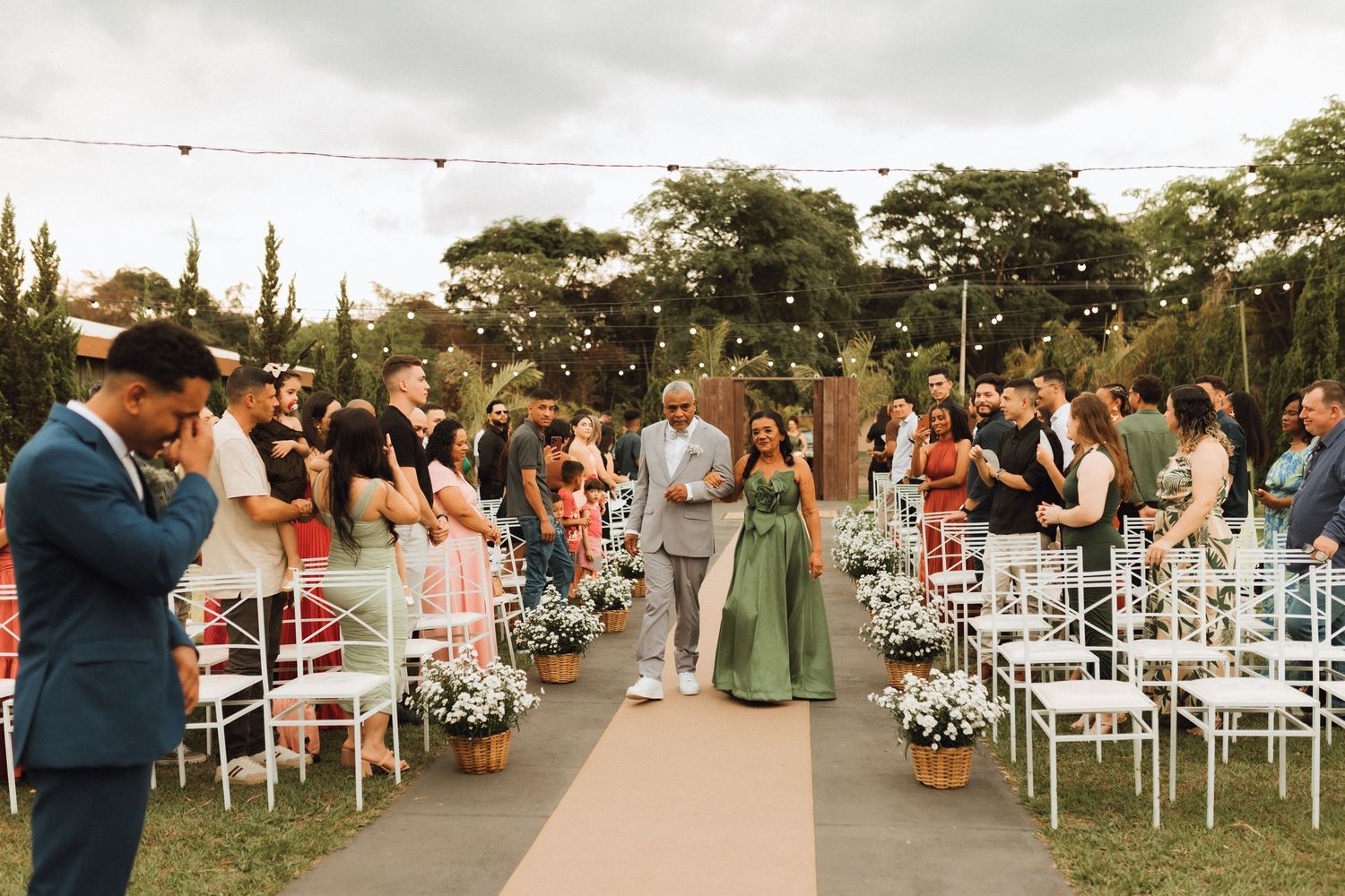 Officiant walks bride in green dress down outdoor wedding aisle lined with white chairs and flowers.