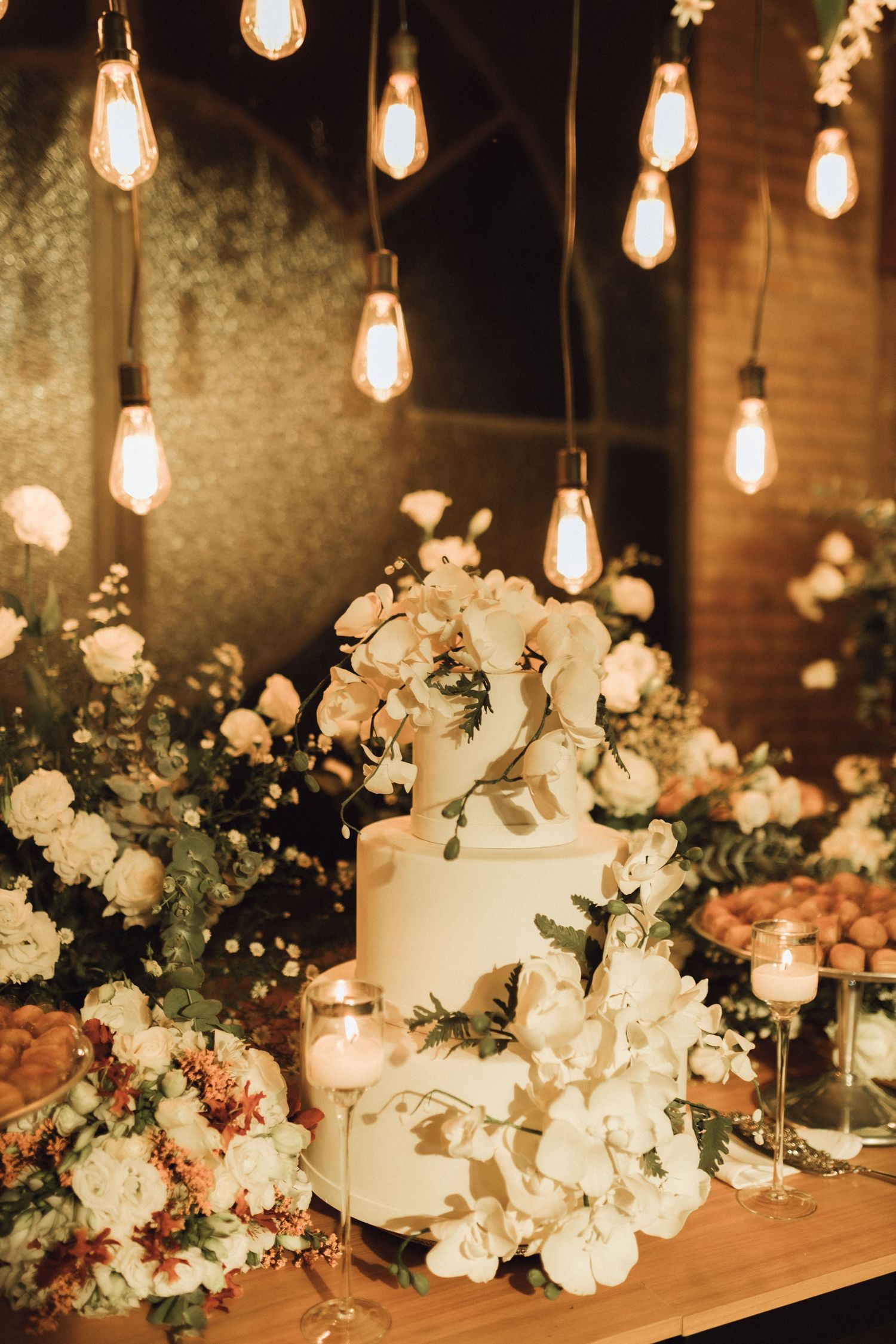 Elegant two-tier white wedding cake adorned with orchids under warm Edison bulb lights.