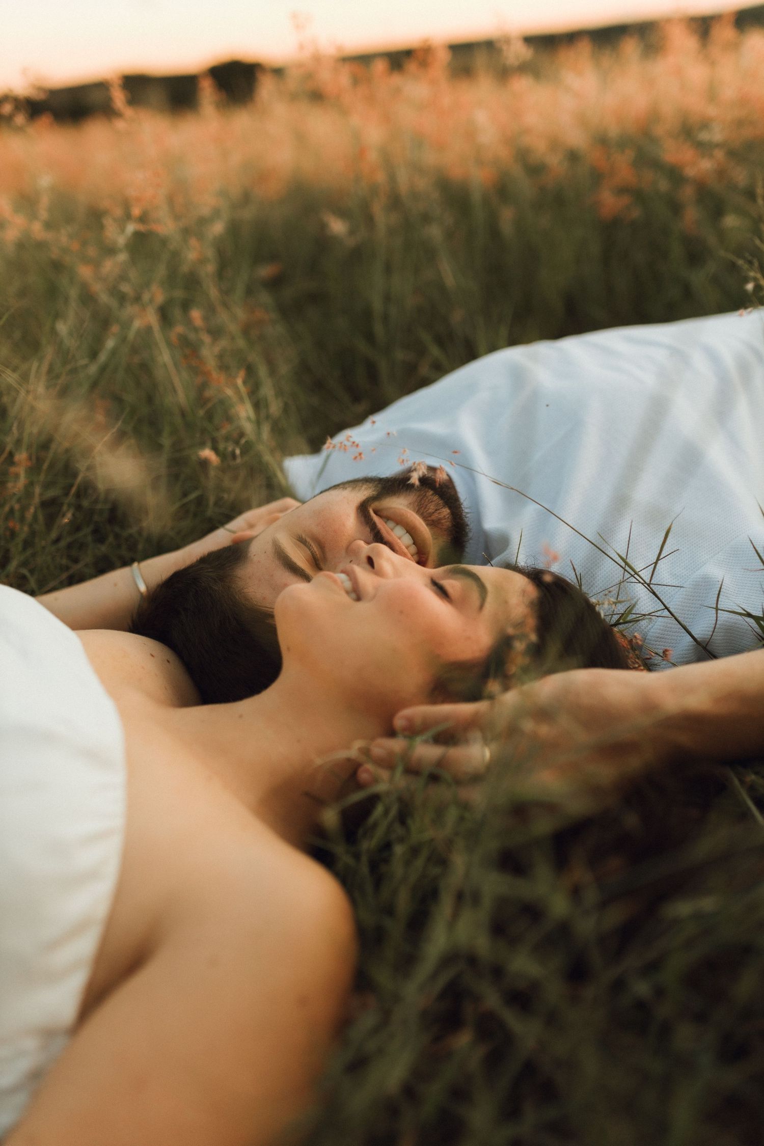 Happy couple lying in tall grass laughing together during a romantic outdoor photo session.