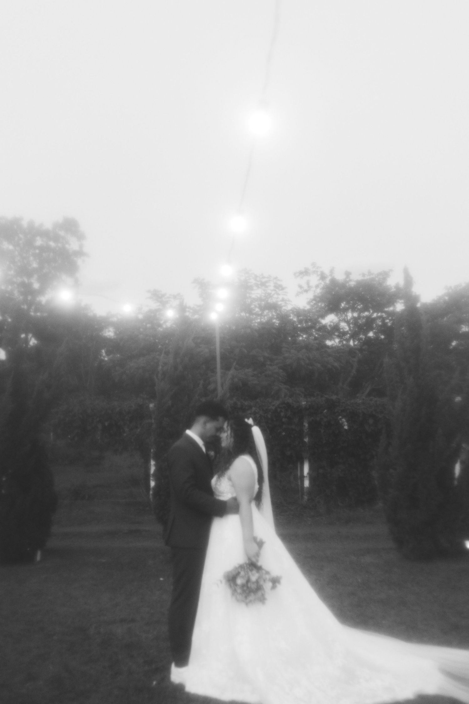 Black and white romantic photo of bride and groom standing under string lights in a garden at dusk.