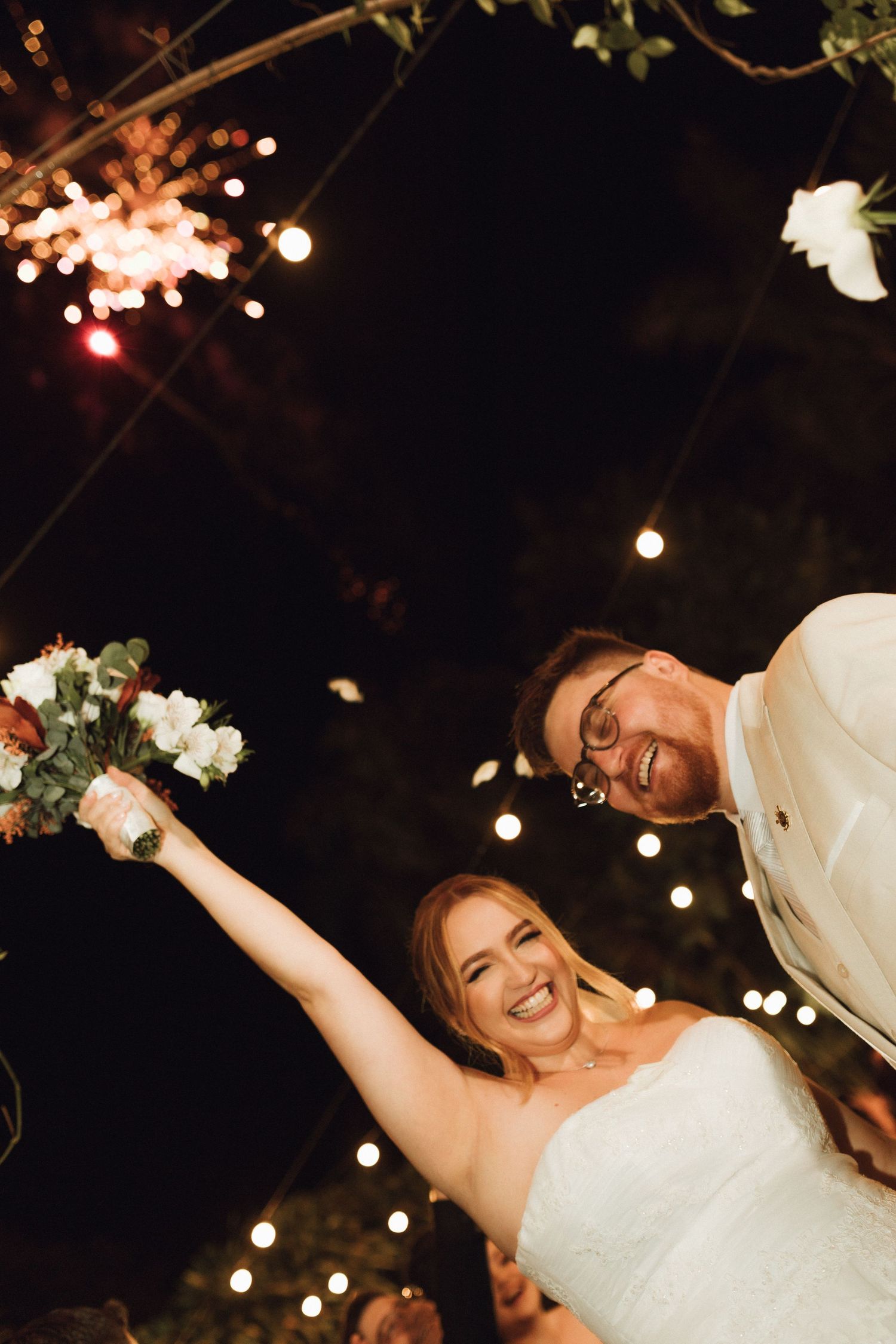 Joyful bride raises bouquet while groom smiles under sparklers and string lights at night.