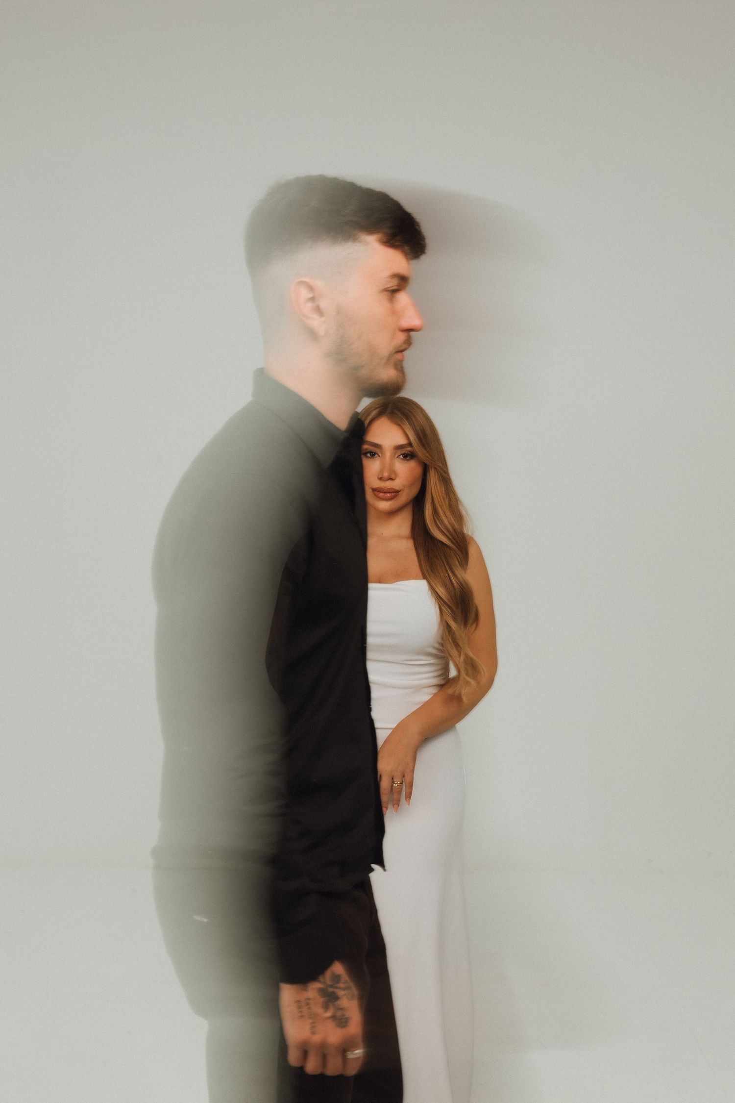 A man in a black shirt and a woman in a white dress pose together against a white wall in a stylish couple portrait.