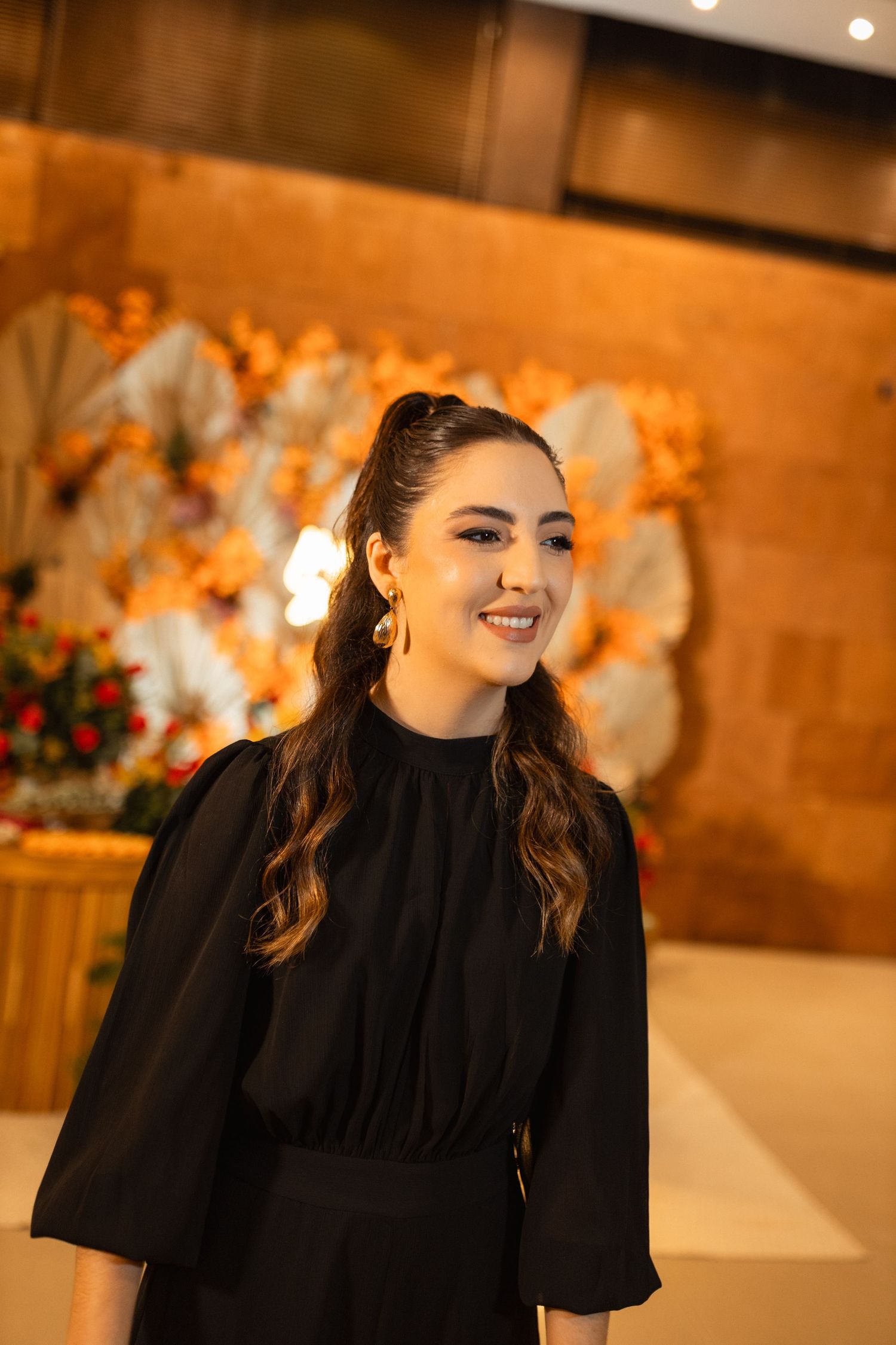 A smiling young woman in a black dress with gold earrings poses in front of a floral backdrop at an event.