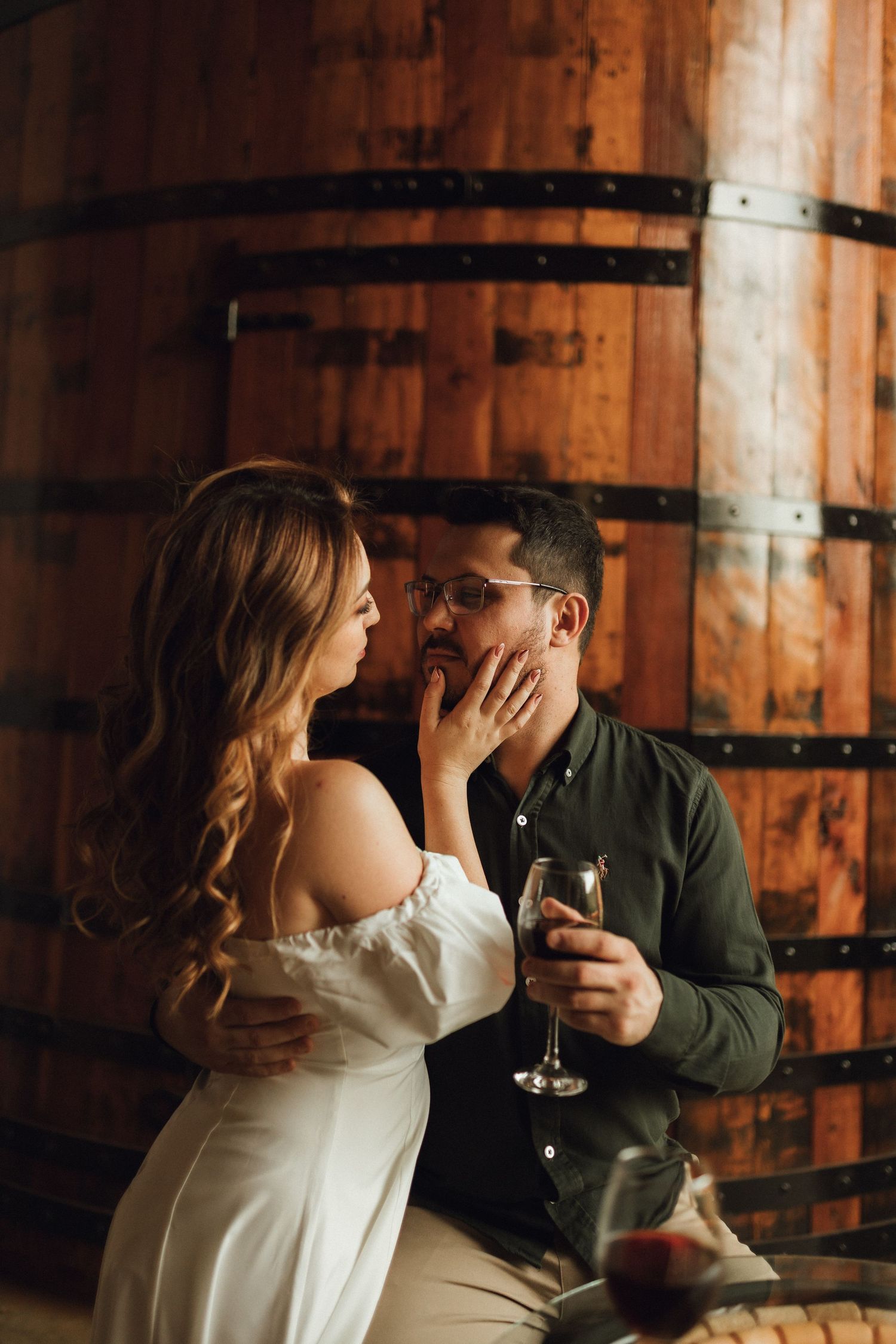 Couple sharing a romantic moment in a winery cellar, woman in white dress touching mans face as he holds a wine glass.