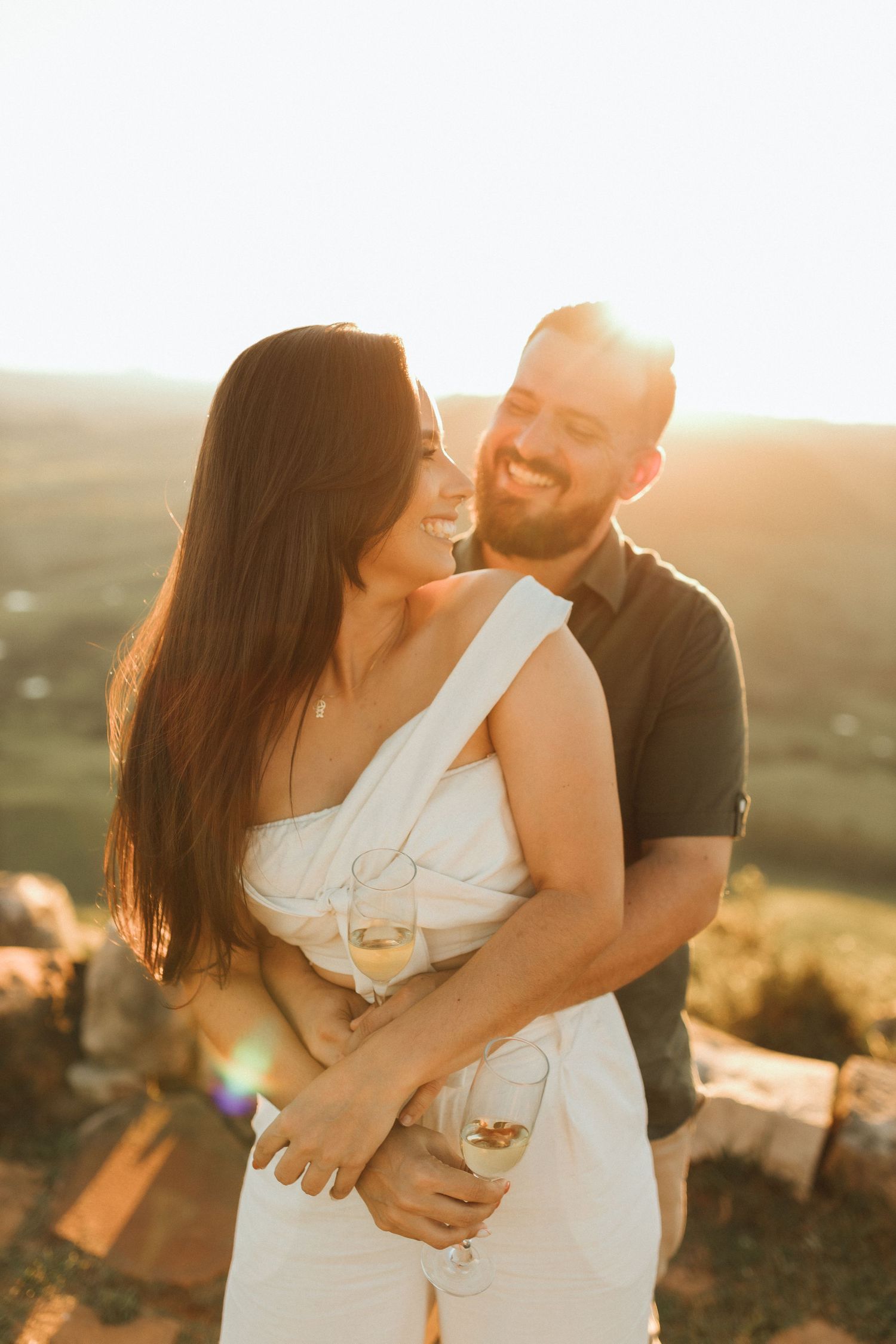 Couple embracing at golden hour sunset, woman in white dress holding wine glass, man smiling warmly outdoors.
