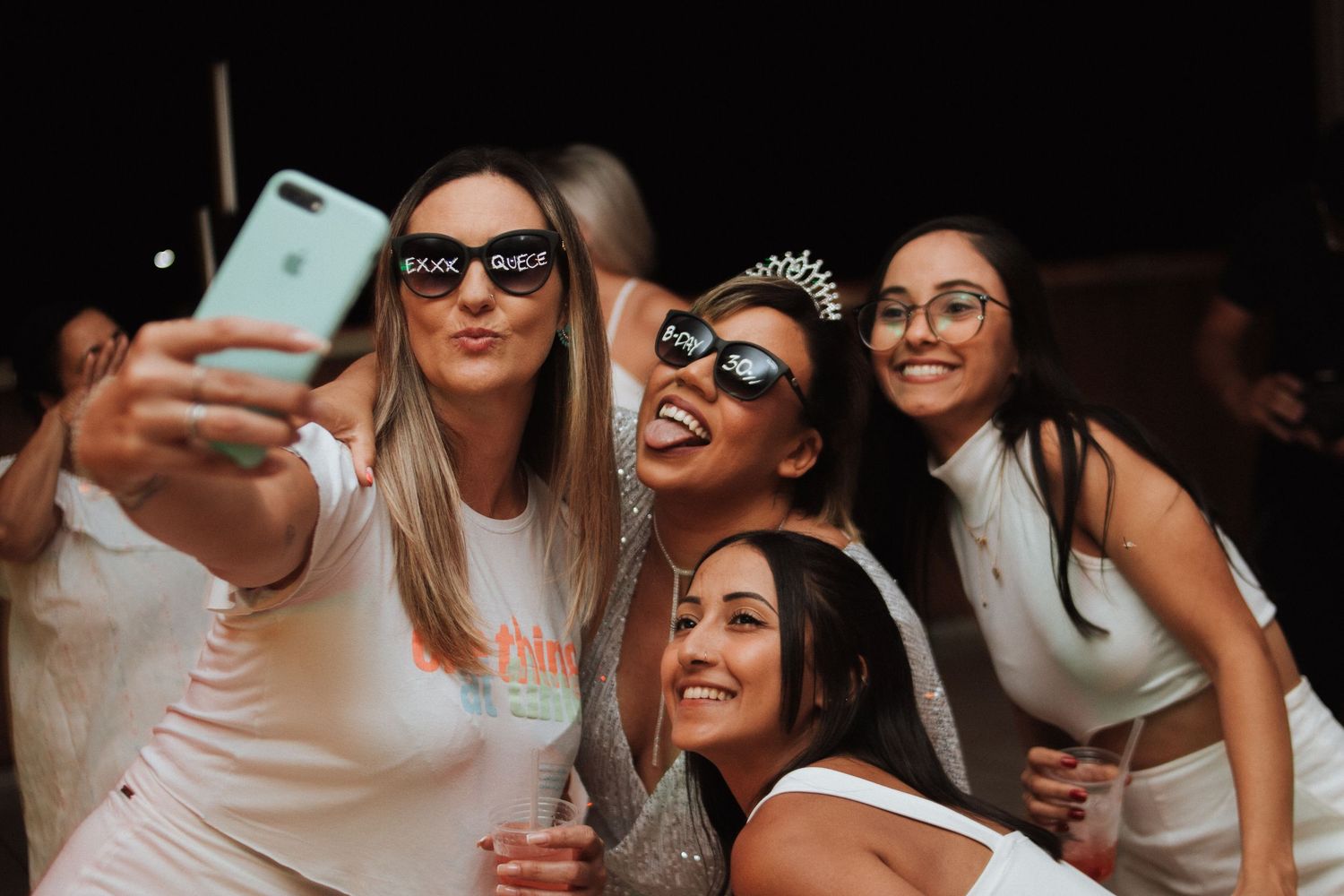 Group of women in white outfits taking a selfie at a party, one wearing a tiara for a bachelorette celebration.