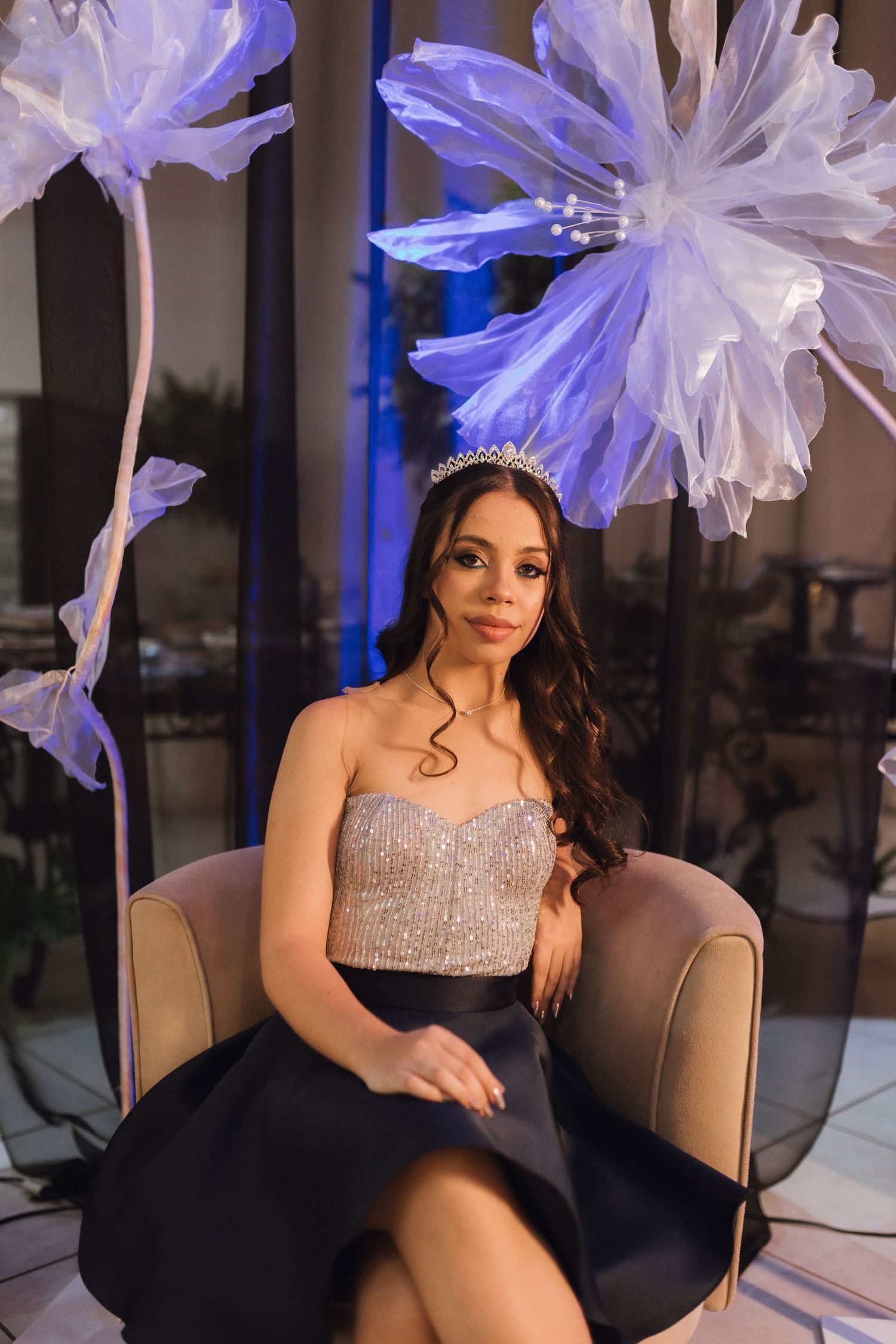 Young woman in sparkly corset top and tiara sits elegantly in chair surrounded by large decorative floral arrangements.