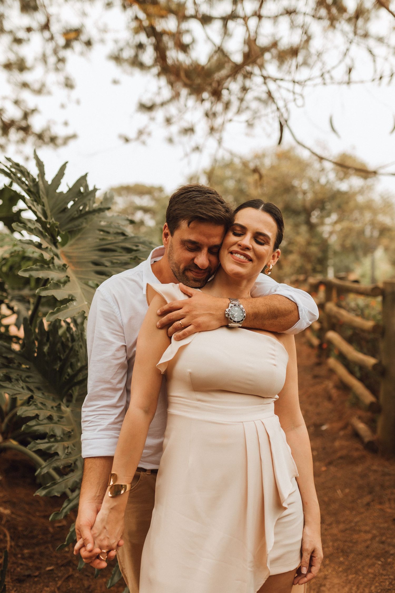 A smiling couple embraces outdoors, the man in a white shirt hugging the woman in a white strapless dress.