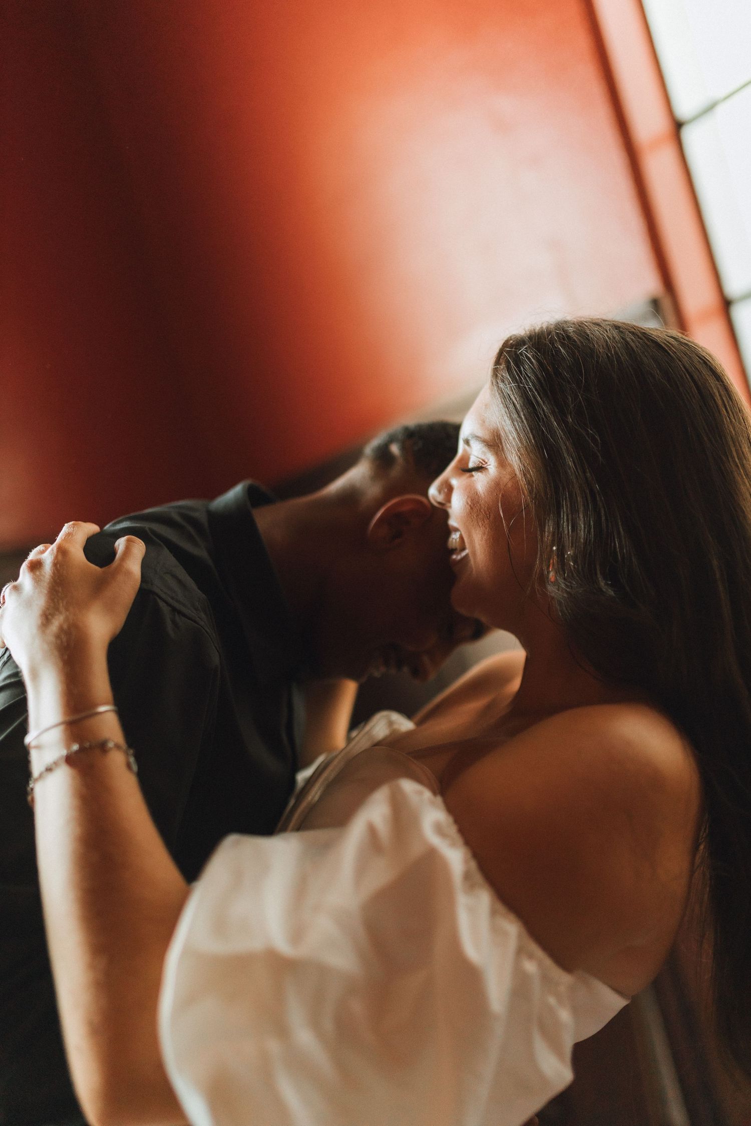Intimate moment of a couple sharing a kiss against a warm red wall in soft romantic lighting.