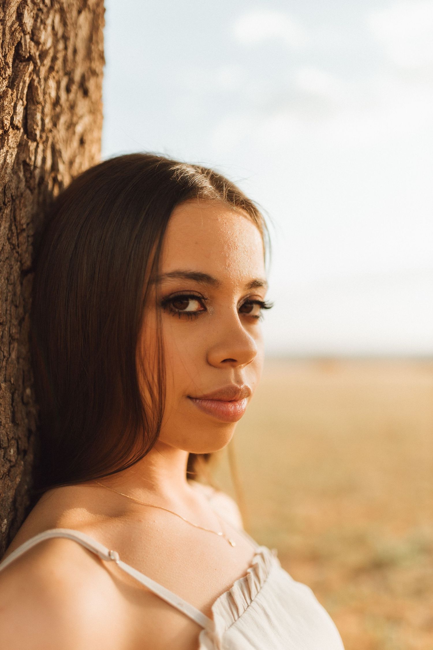 Young woman with long dark hair leaning against a tree in a golden field, wearing a white dress.