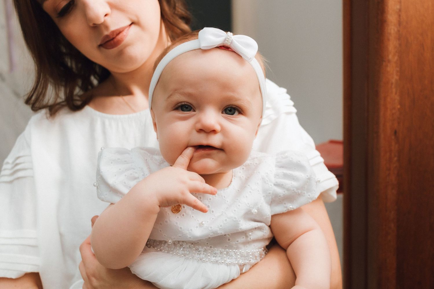 A mother in white dress holds her adorable baby girl wearing a white outfit and bow headband, smiling cutely.