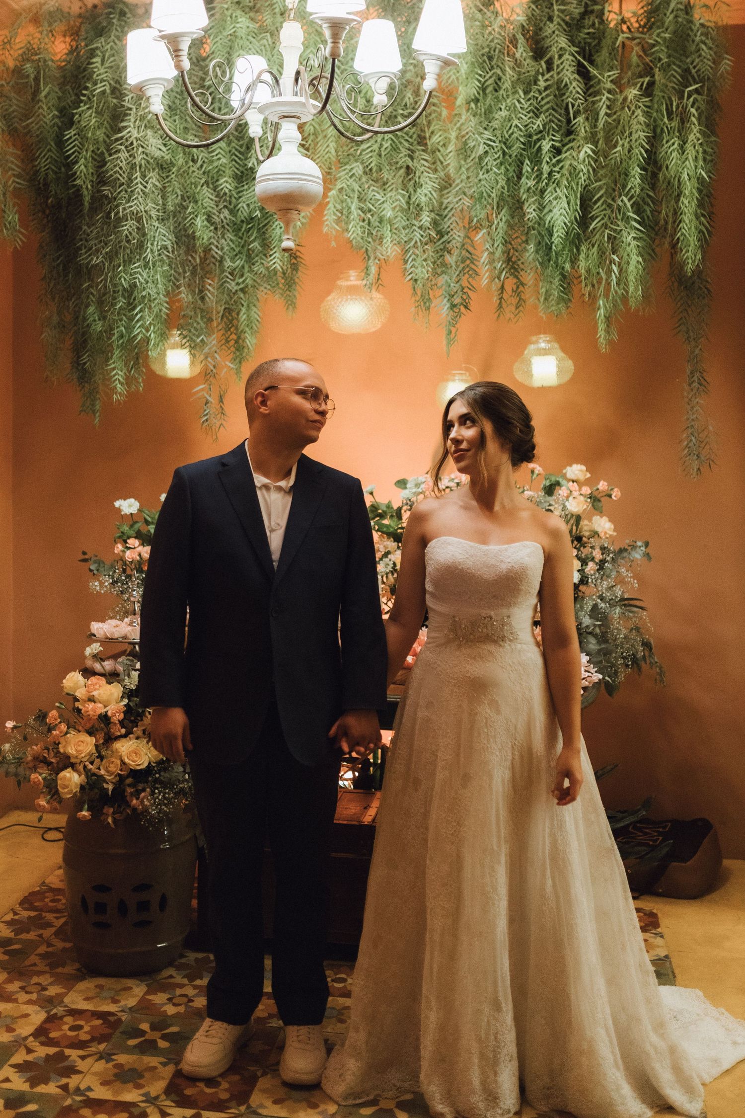 Newlyweds hold hands under hanging greenery chandelier surrounded by floral arrangements at reception.