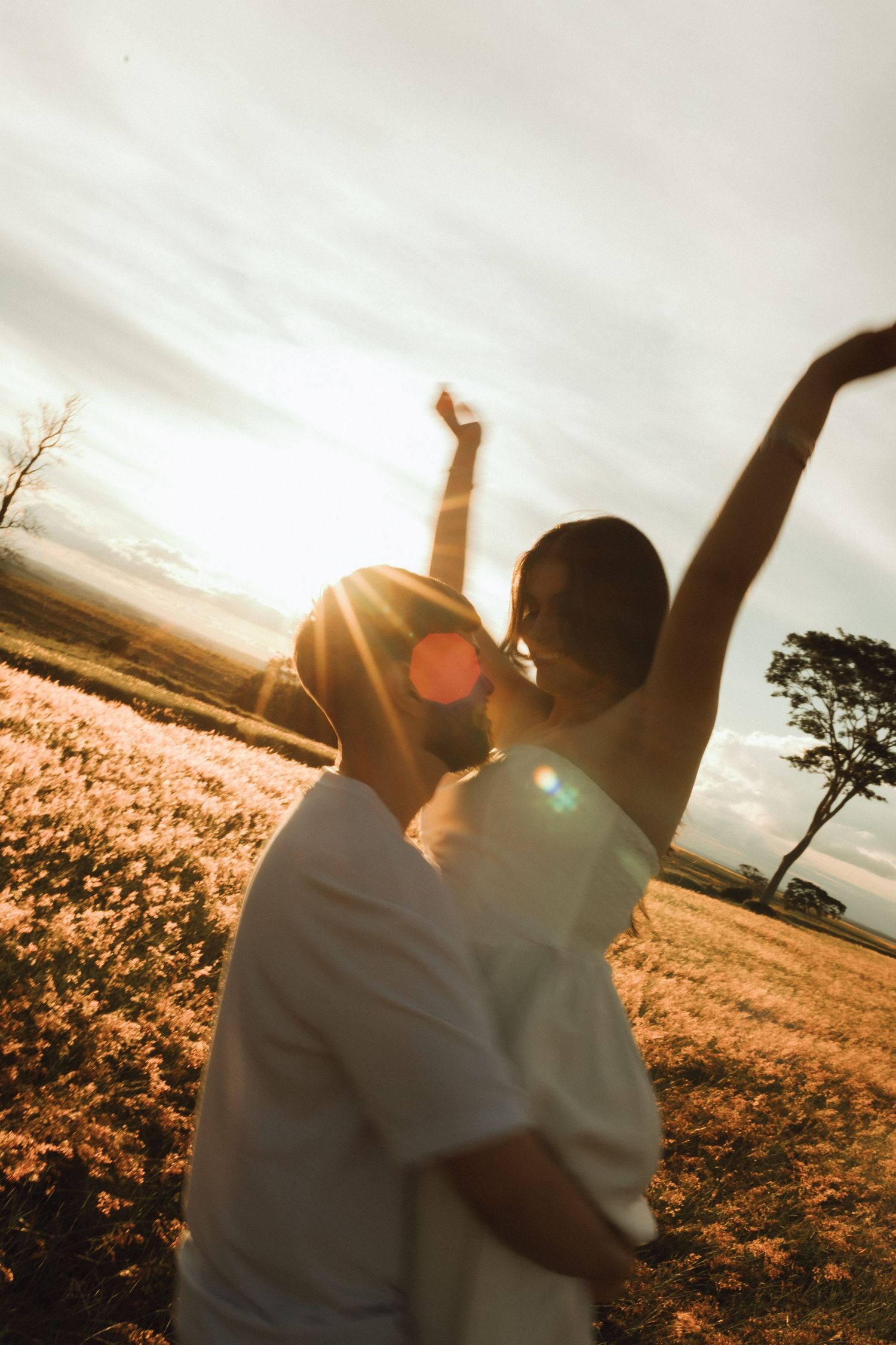 Couple embracing at golden hour sunset in a field, woman raising arms joyfully with lens flare.