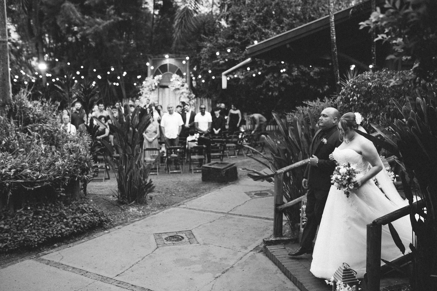 Black and white photo of bride and escort entering outdoor garden wedding ceremony at night.