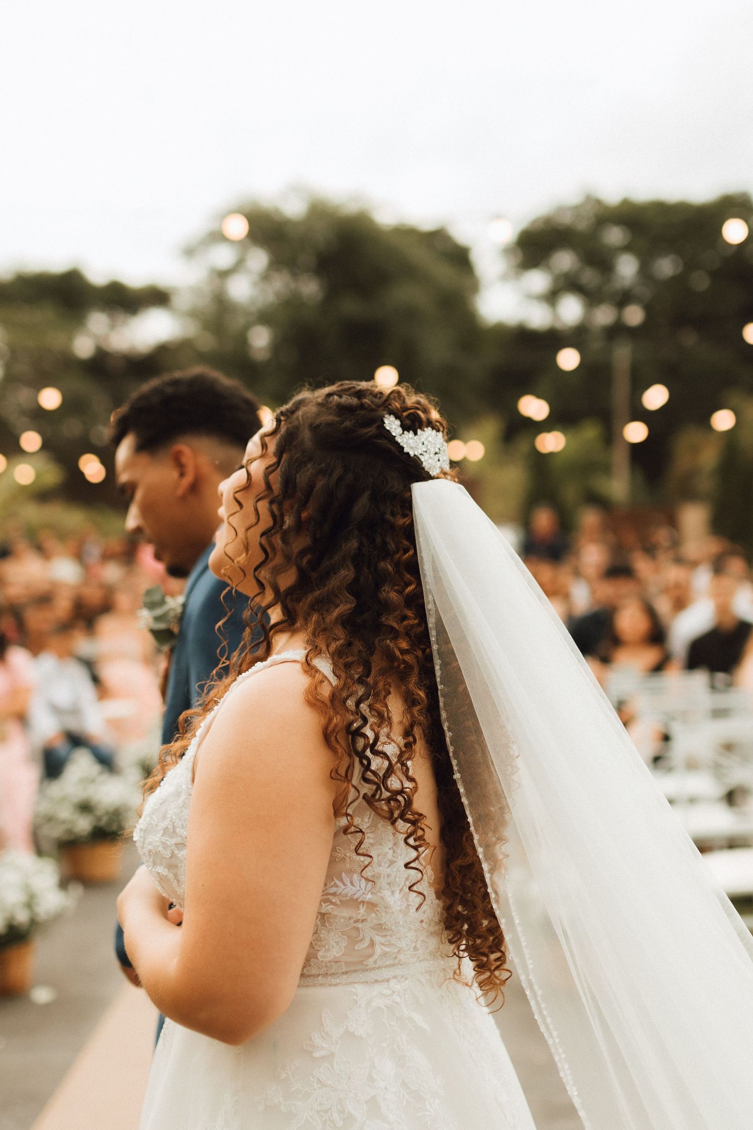 Bride with curly hair and floral hair accessory stands at outdoor wedding ceremony with string lights.
