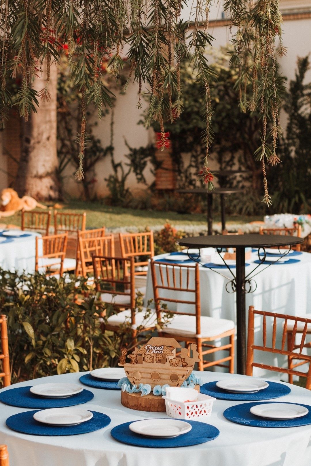 Elegant outdoor wedding reception with blue placemats, white plates, wooden chairs, and hanging greenery decor.