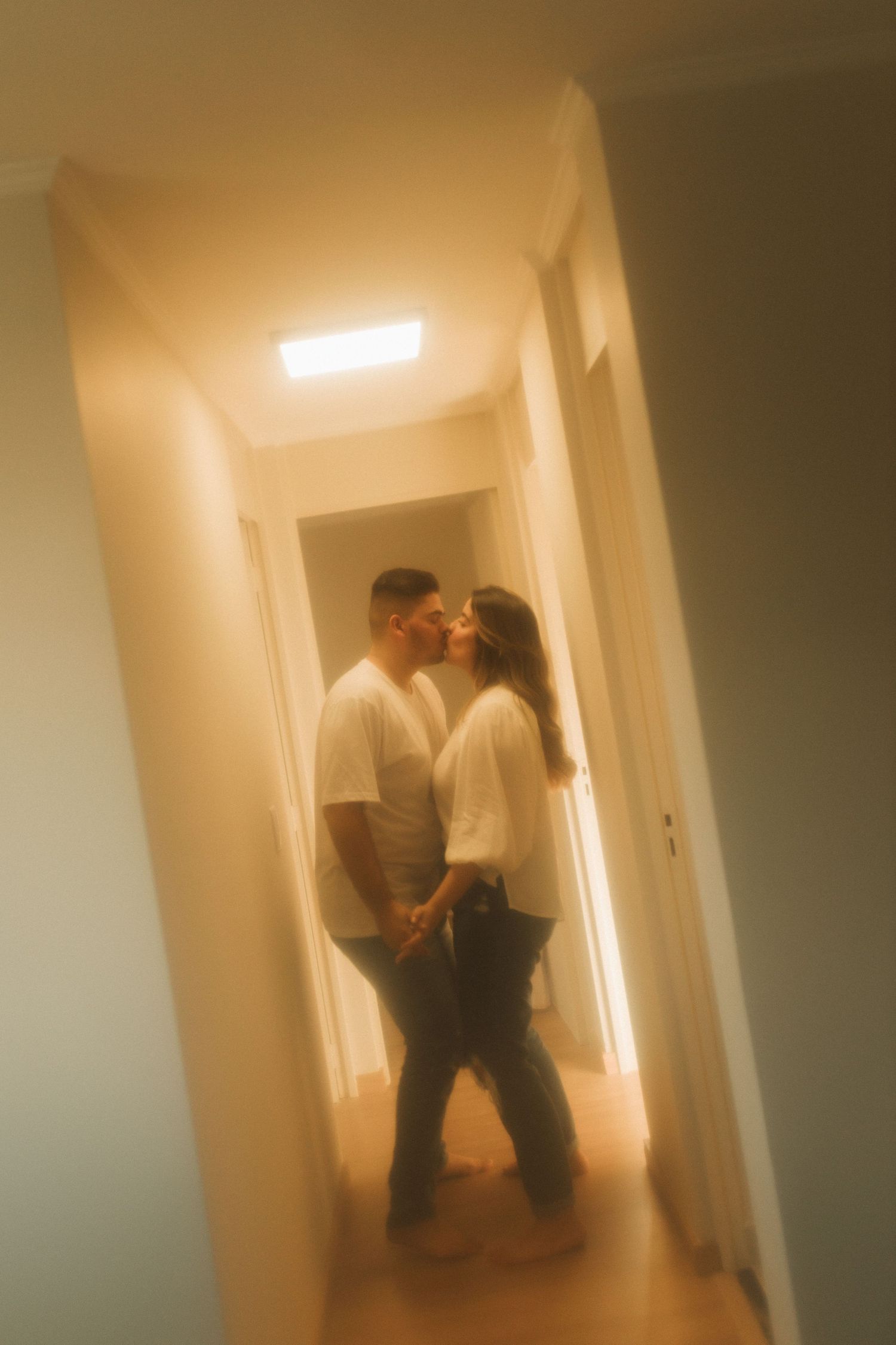 Couple sharing a romantic kiss in a warmly lit hallway, both wearing white shirts and jeans.