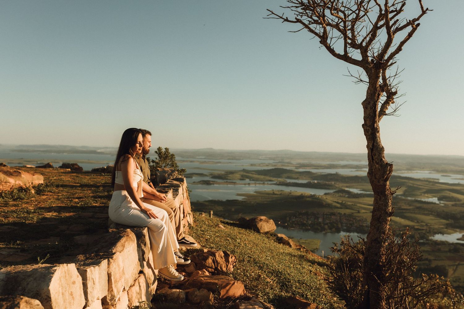 Couple sitting on stone wall at golden hour overlooking scenic landscape with bare tree and distant water views.