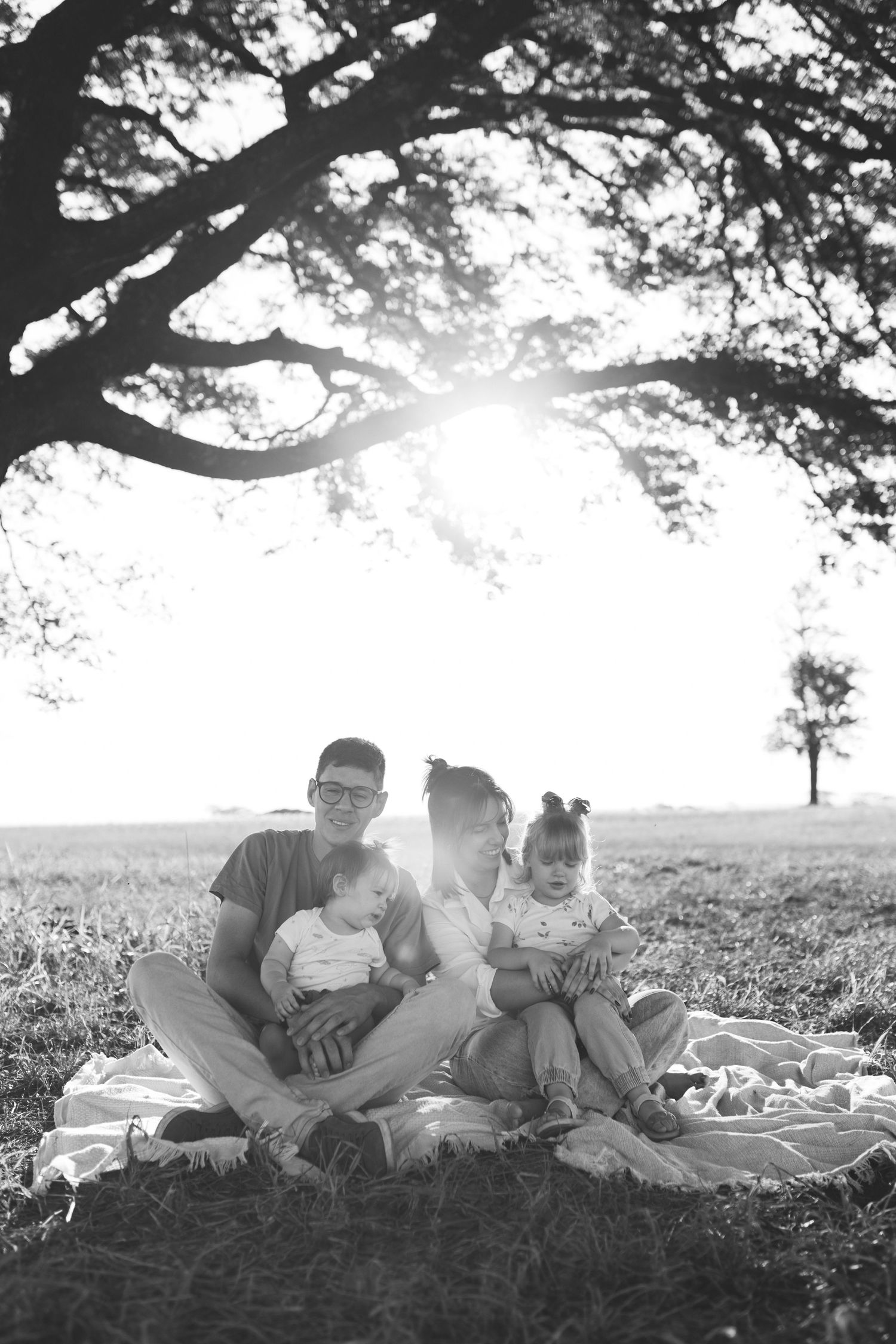 Black and white family portrait sitting on blanket under large tree in field.